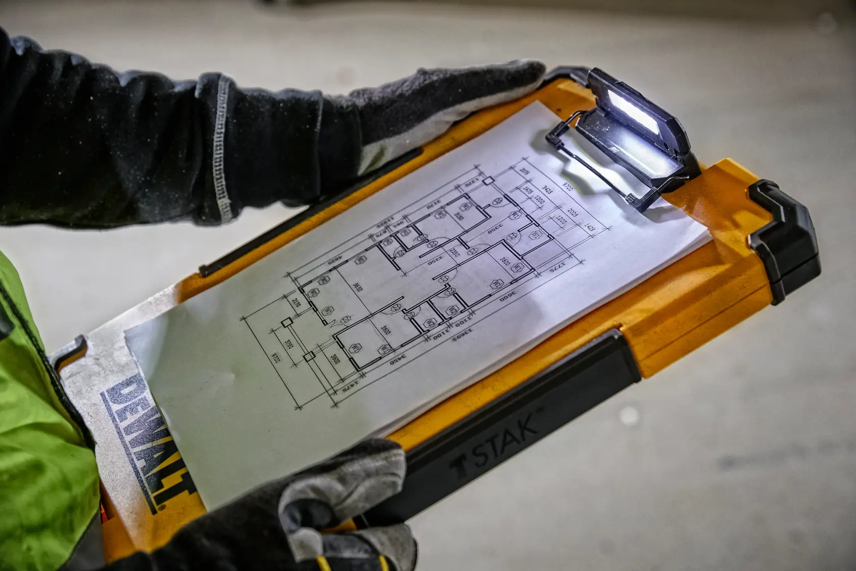 A person wearing work gloves holds a DEWALT TSTAK clipboard with an architectural blueprint attached. The clipboard features an integrated LED light to illuminate the document.