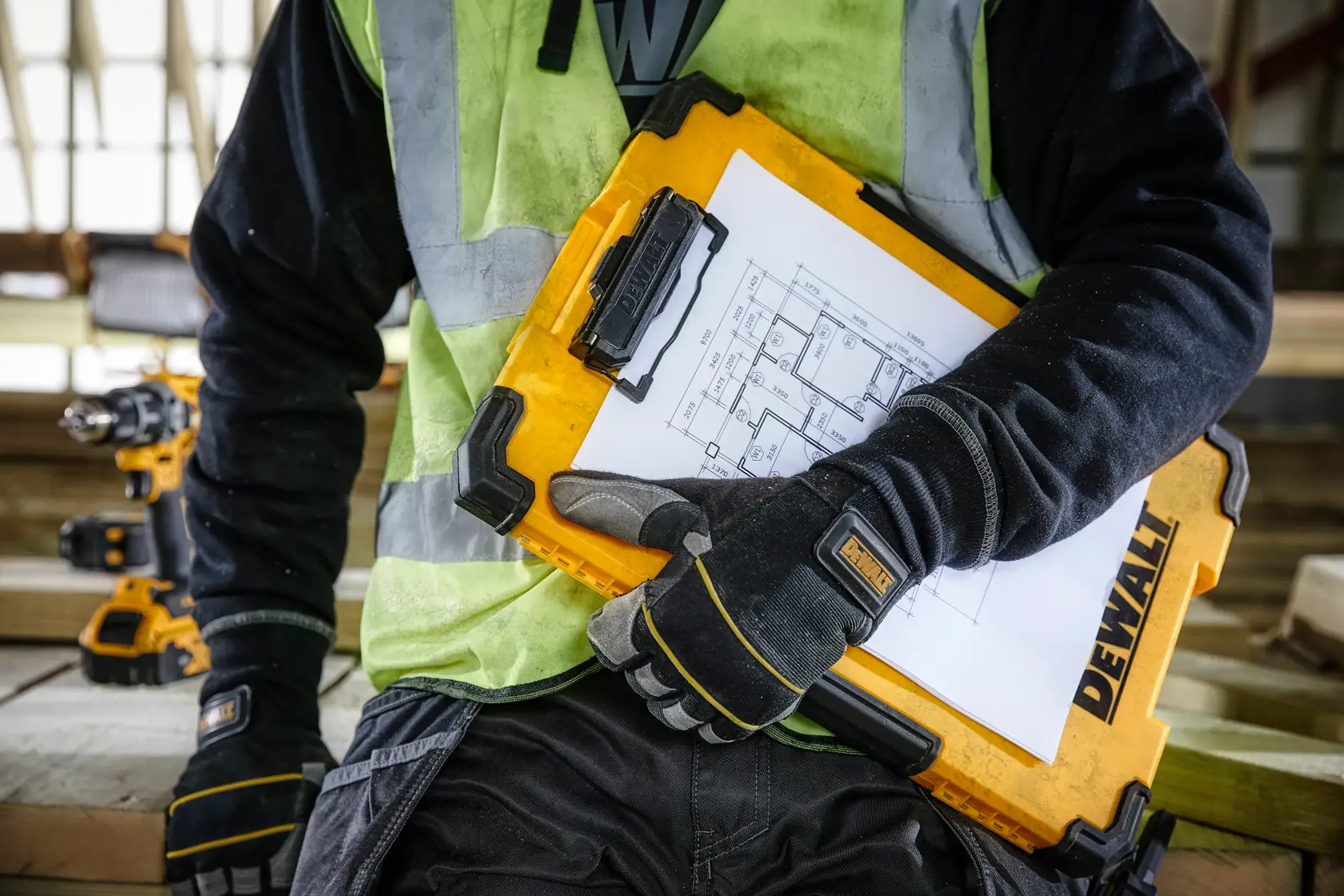 A person wearing work gloves and a safety vest holds a yellow DEWALT DWST17818 case containing construction blueprints, with power tools visible in the background.