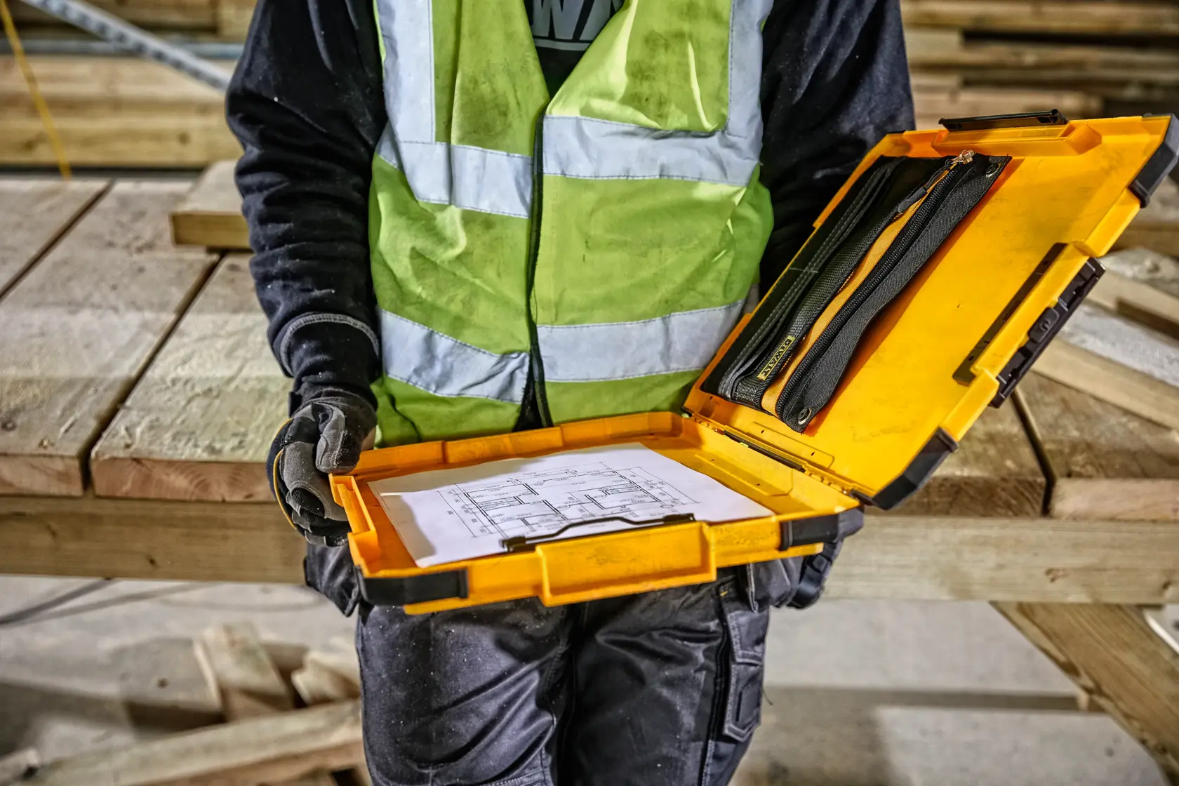 A person wearing a high-visibility vest and gloves is holding an open yellow DEWALT case containing work documents and some tools, with a wooden construction site in the background.