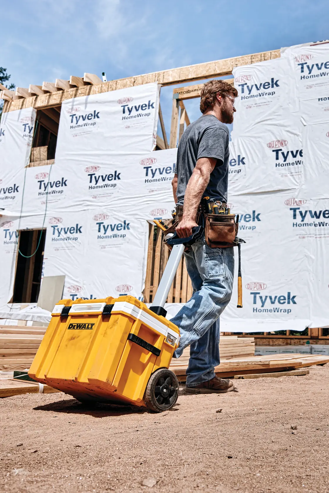 A person at a construction site is pulling a yellow DEWALT rolling toolbox on dirt, in front of a building partially wrapped with Tyvek HomeWrap. The person is wearing a tool belt and jeans.