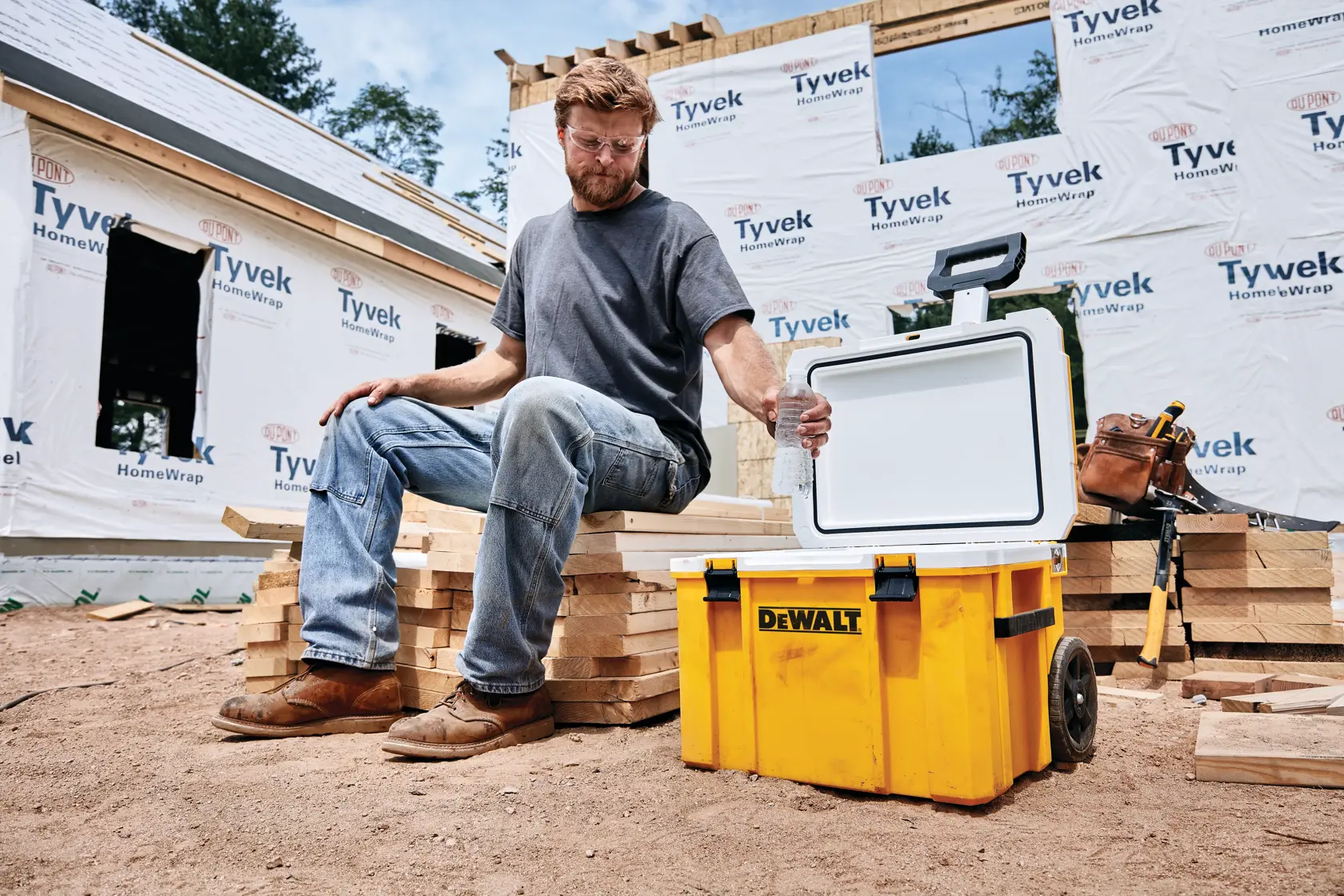 A person sitting on stacked wooden planks at a construction site with a yellow DEWALT rolling cooler with an open lid beside them. Partially built house and construction materials are visible in the background.