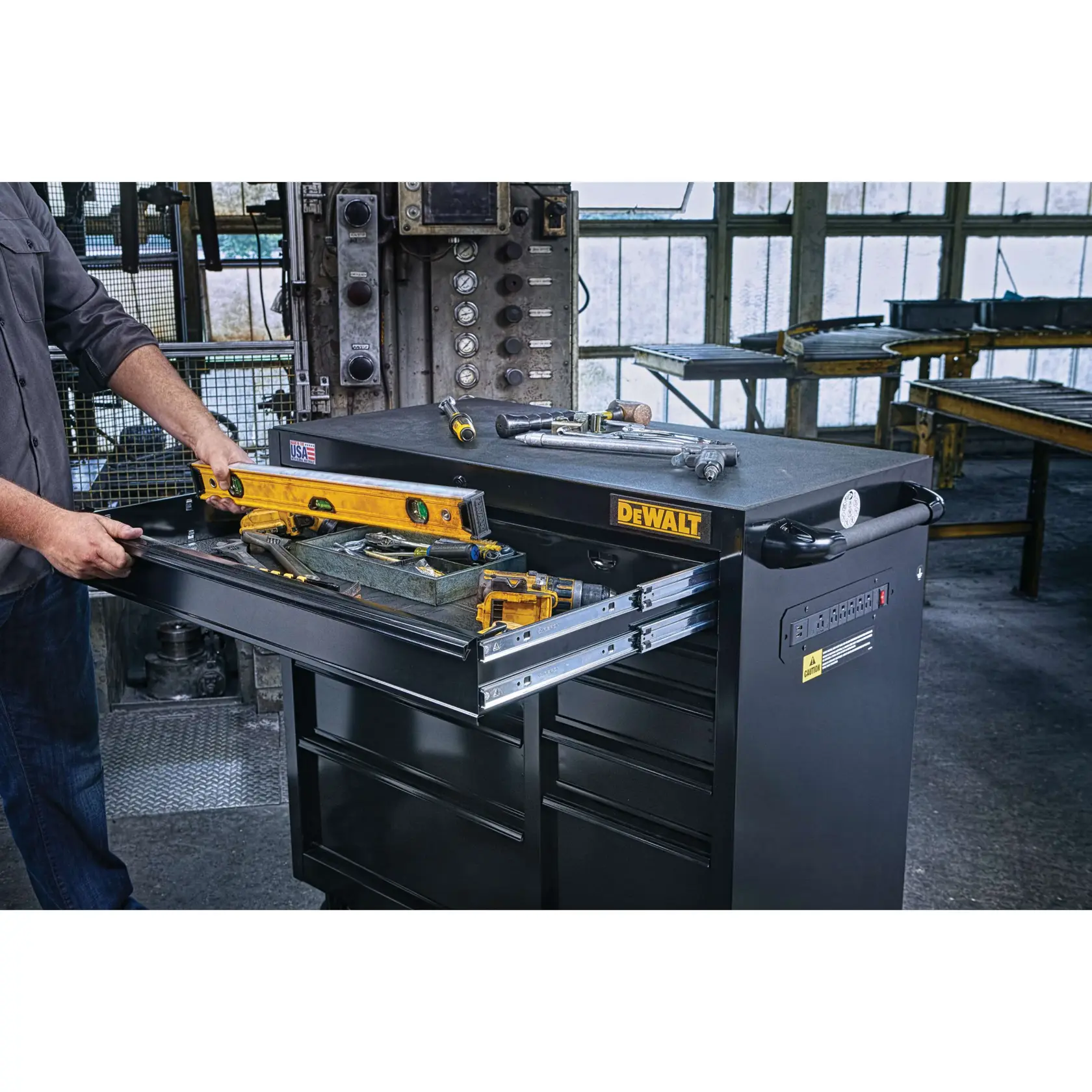 A person opens a drawer of a DEWALT tool chest filled with various hand tools, including a level and tape measure, set in an industrial workshop environment.