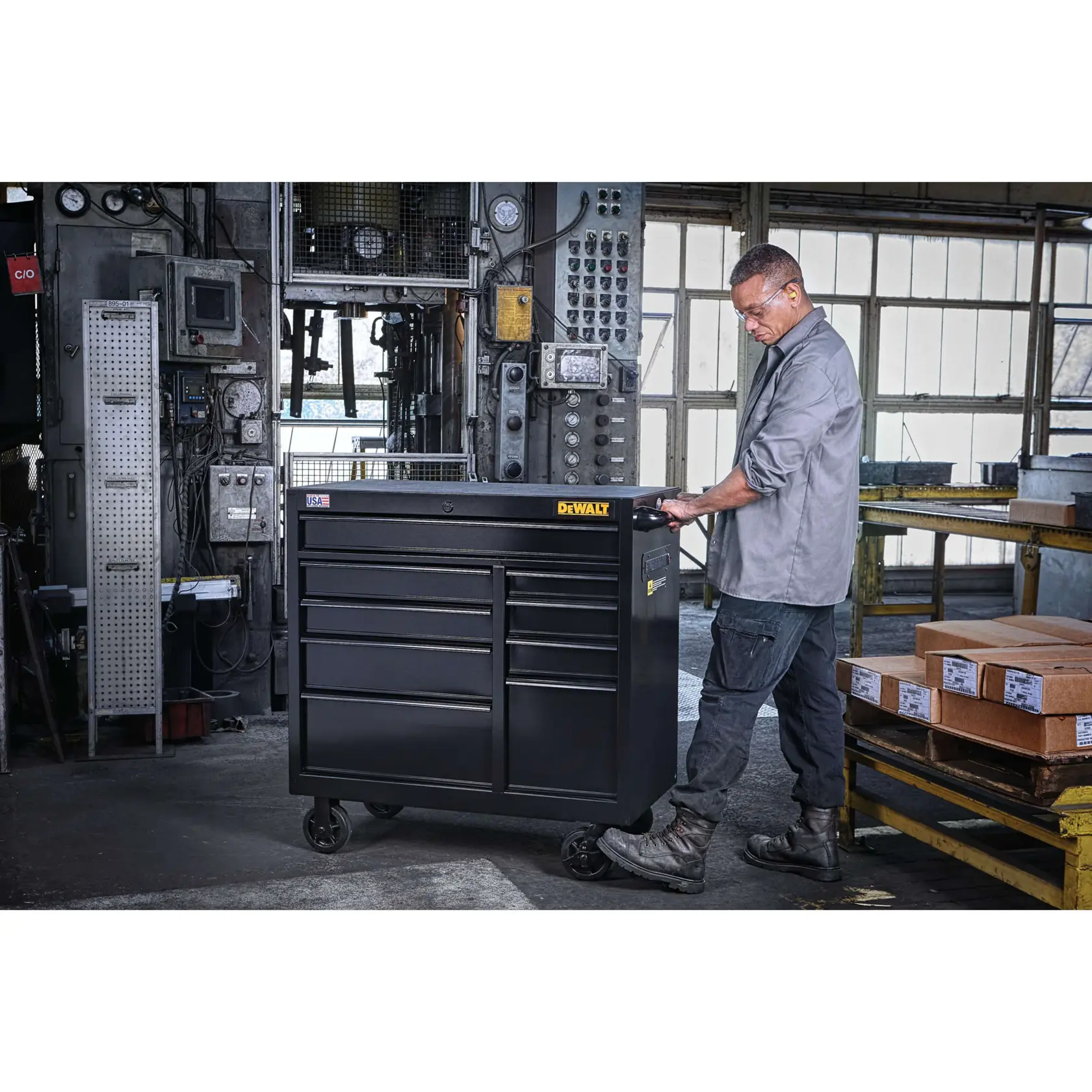A person in a workshop setting moves a black DEWALT rolling tool chest with multiple drawers and wheels. Industrial equipment and packed boxes are visible in the background.
