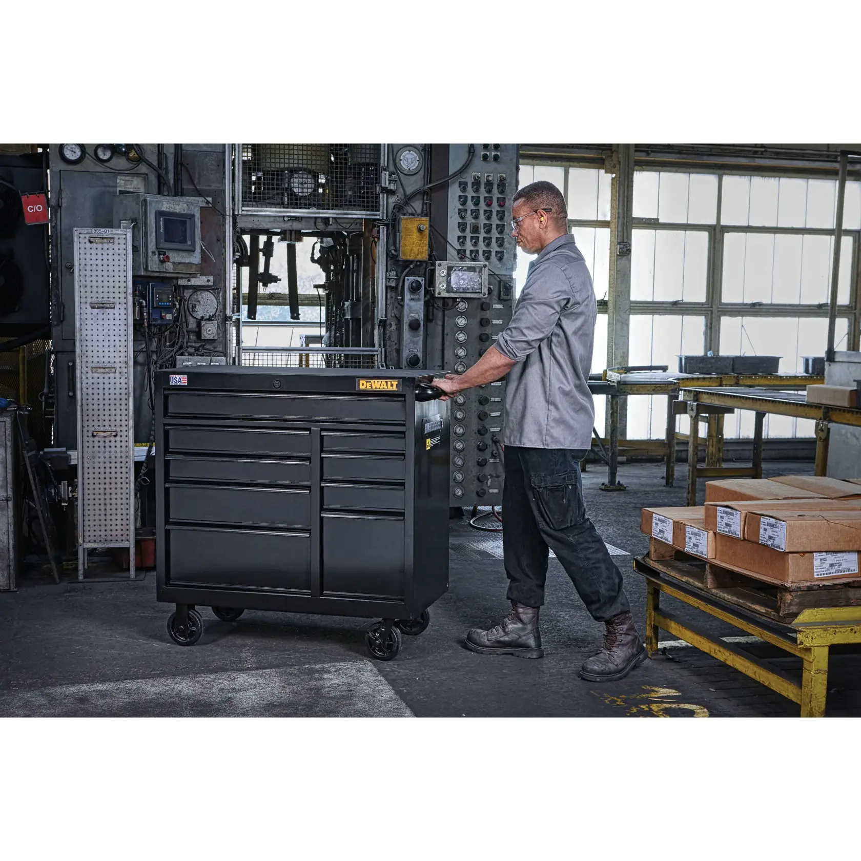 A person pushing a DEWALT DWST24192 rolling tool chest with multiple drawers in an industrial workshop setting. The tool chest is black and has wheels for mobility.