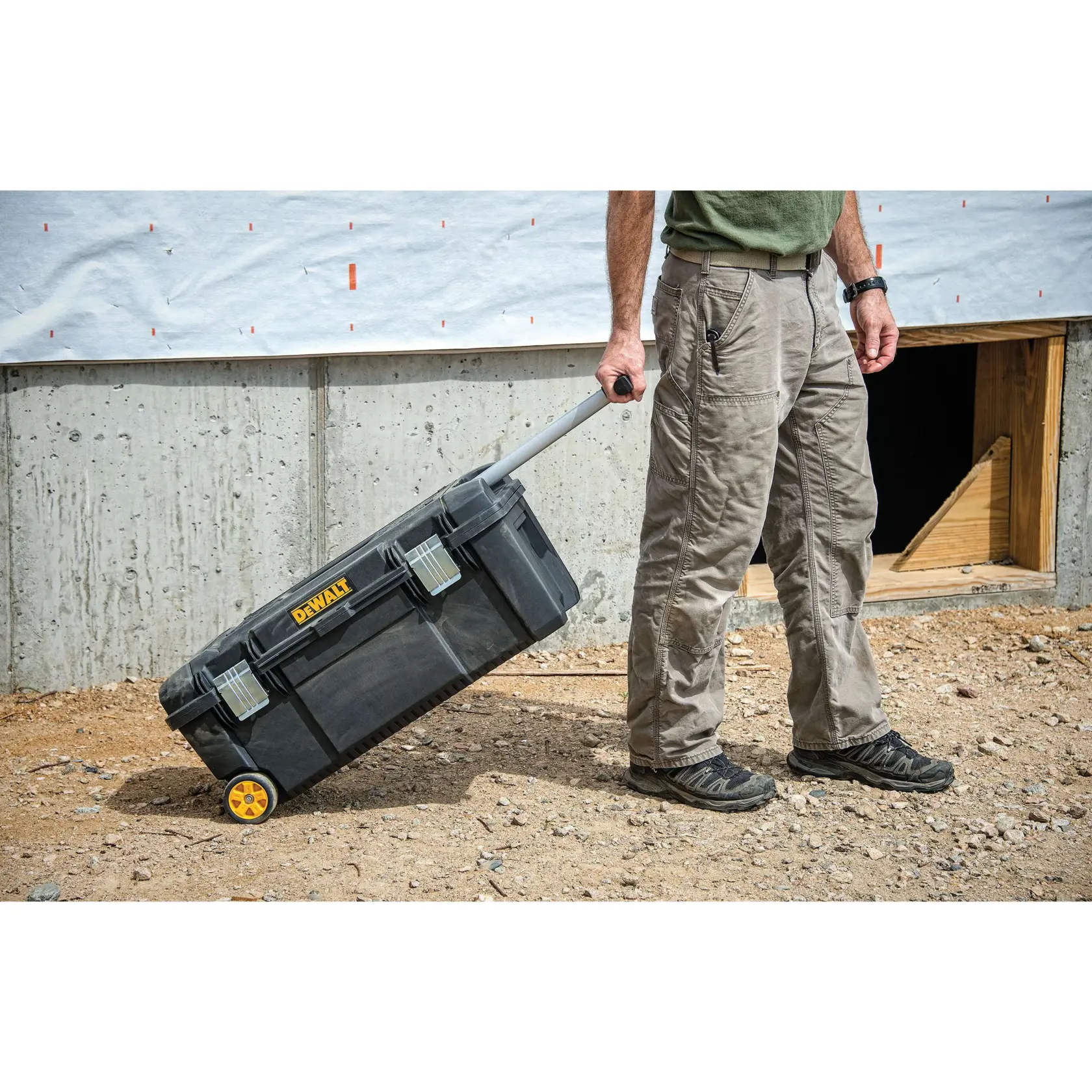 A person pulls a black DEWALT rolling toolbox with yellow wheels and a telescopic handle on a construction site with dirt and a concrete wall.