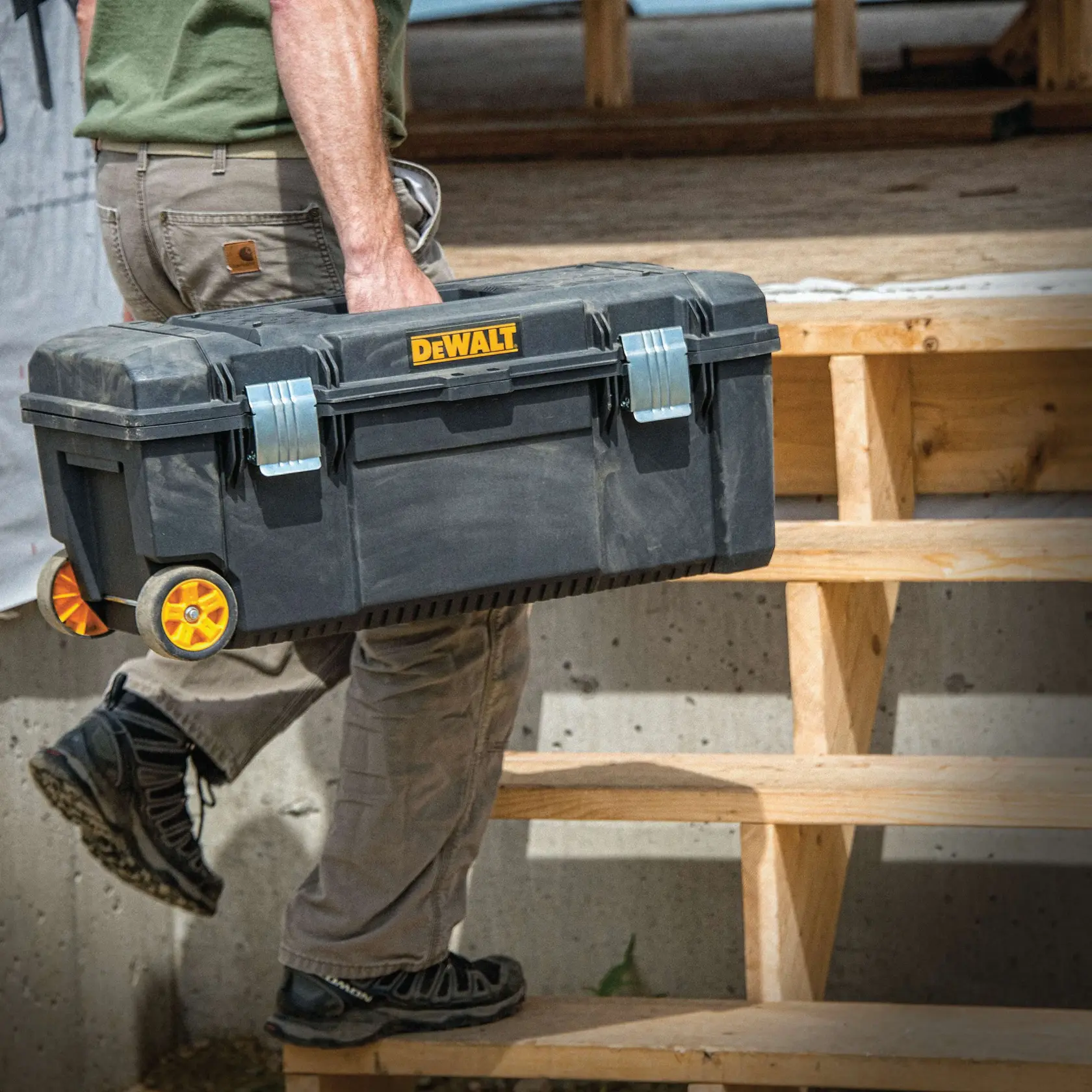 A person carrying a DEWALT rolling toolbox up wooden steps at a construction site.