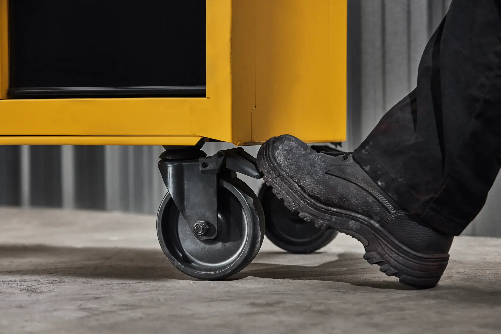 Close-up of a person wearing black work shoes using their foot to engage the brake on the caster wheel of a yellow DEWALT tool storage cart.