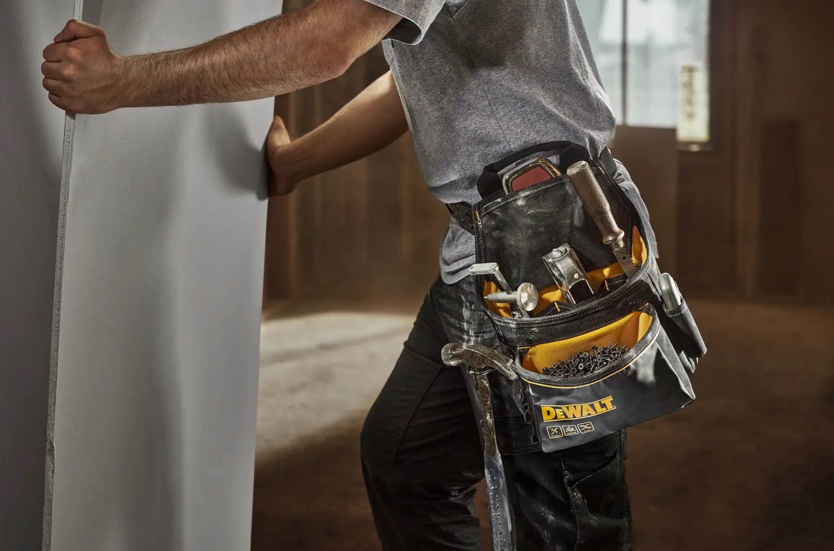 A person wearing a DEWALT branded tool belt filled with various tools and hardware, standing indoors and holding a large panel, dressed in work clothes.