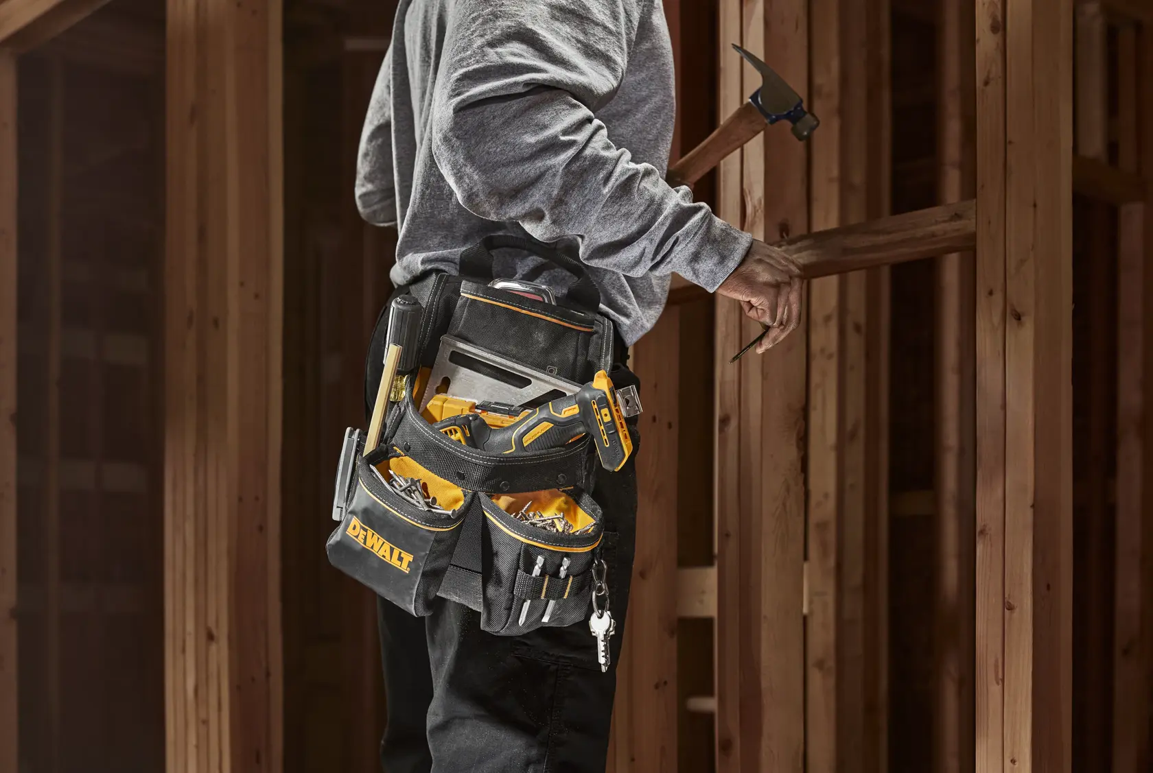 Person wearing a DEWALT tool belt loaded with various hand tools and construction accessories, standing at a wooden construction site and holding a hammer.