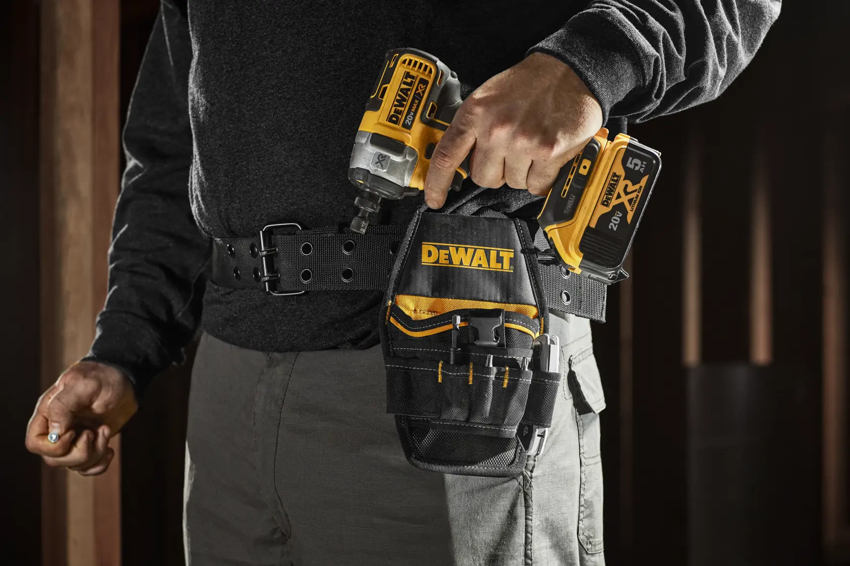 A person wearing a tool belt with a DEWALT branded pouch, holding a DEWALT cordless power drill. The pouch holds various hand tools and the person is dressed in work attire, standing indoors.