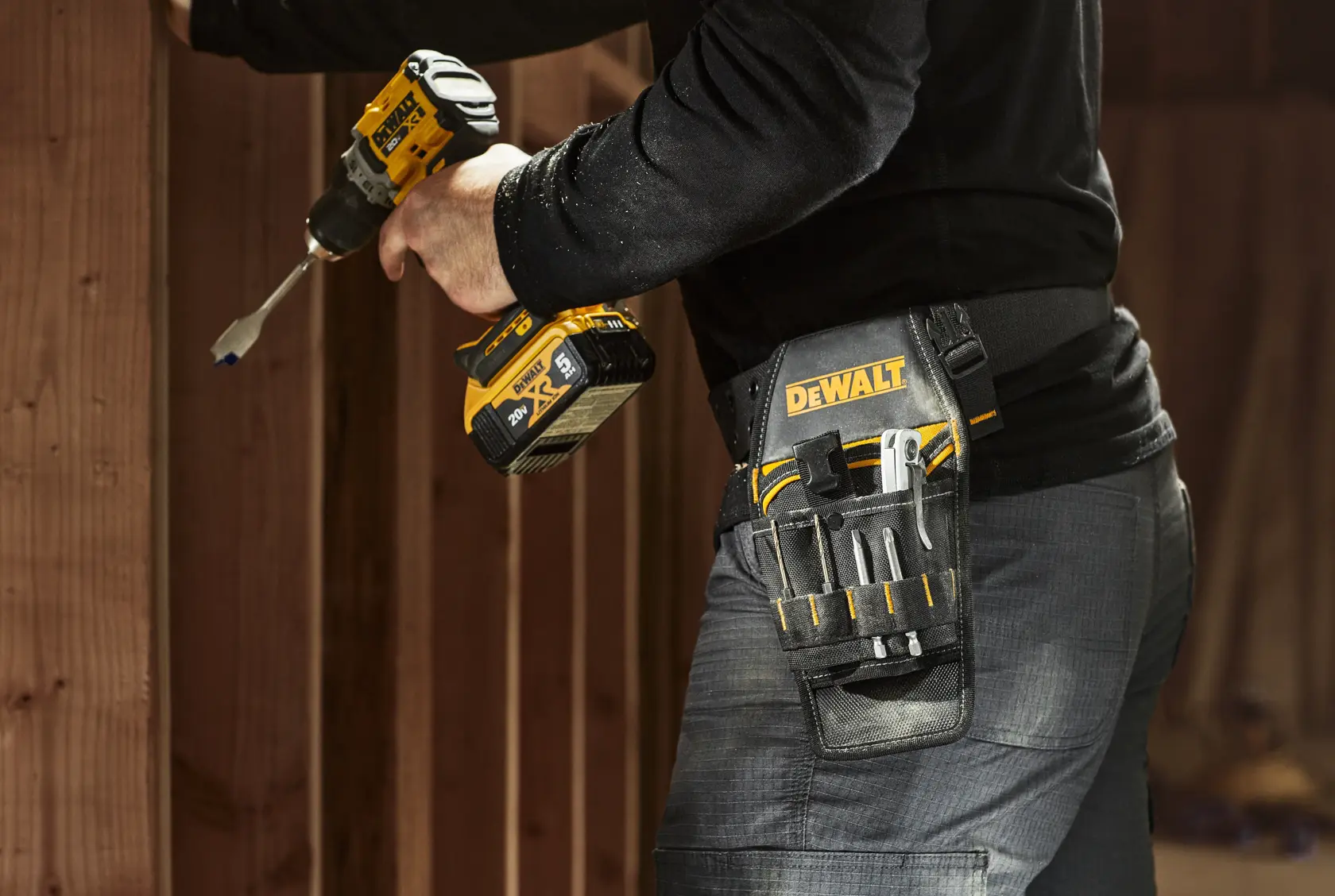 A person is using a DEWALT cordless power drill next to wooden beams. The person is wearing a DEWALT branded tool belt pouch that holds several tools such as screwdrivers and a utility knife.