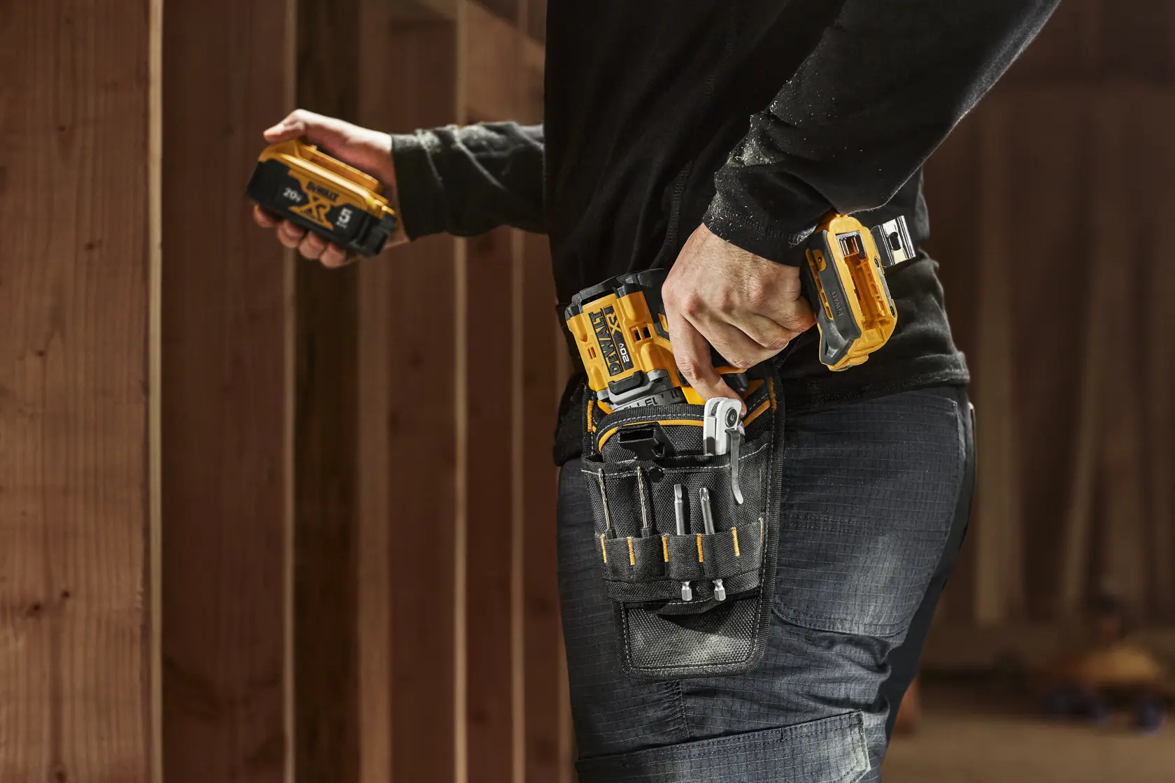A person wearing a tool belt with various tools and holding a DEWALT cordless power drill in a wooden construction setting.