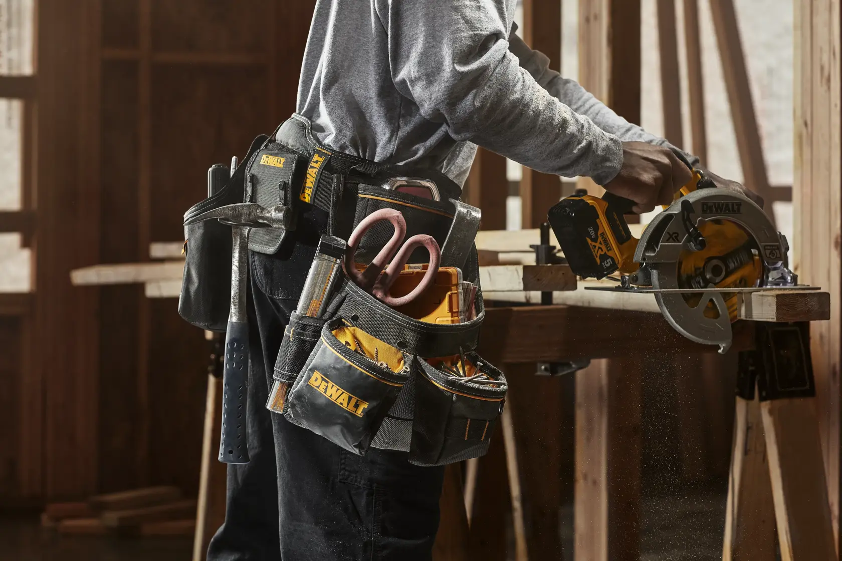 A person wearing a DEWALT tool belt with various tools, including a hammer, measuring tape, scissors, and other hand tools, is using a DEWALT circular saw to cut wood in a construction setting.