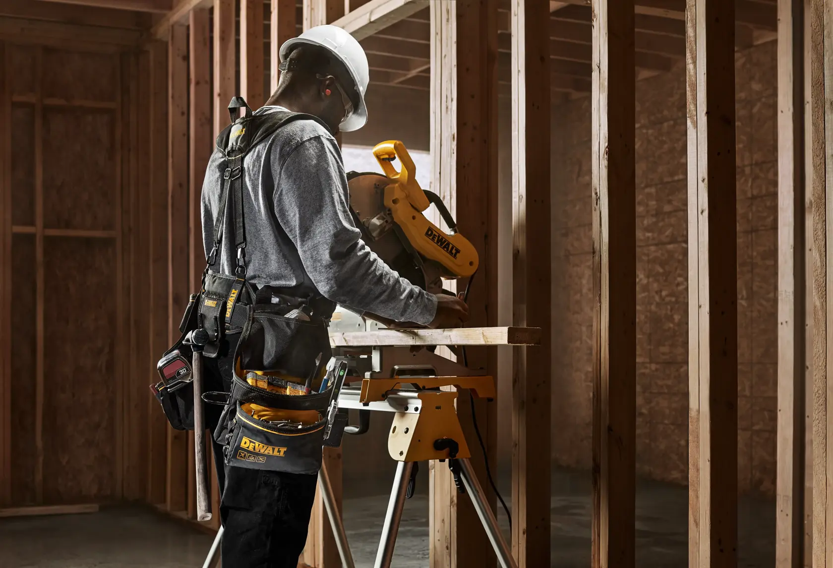 A person wearing a hard hat and tool belt uses a DEWALT miter saw in a wood-framed construction site.