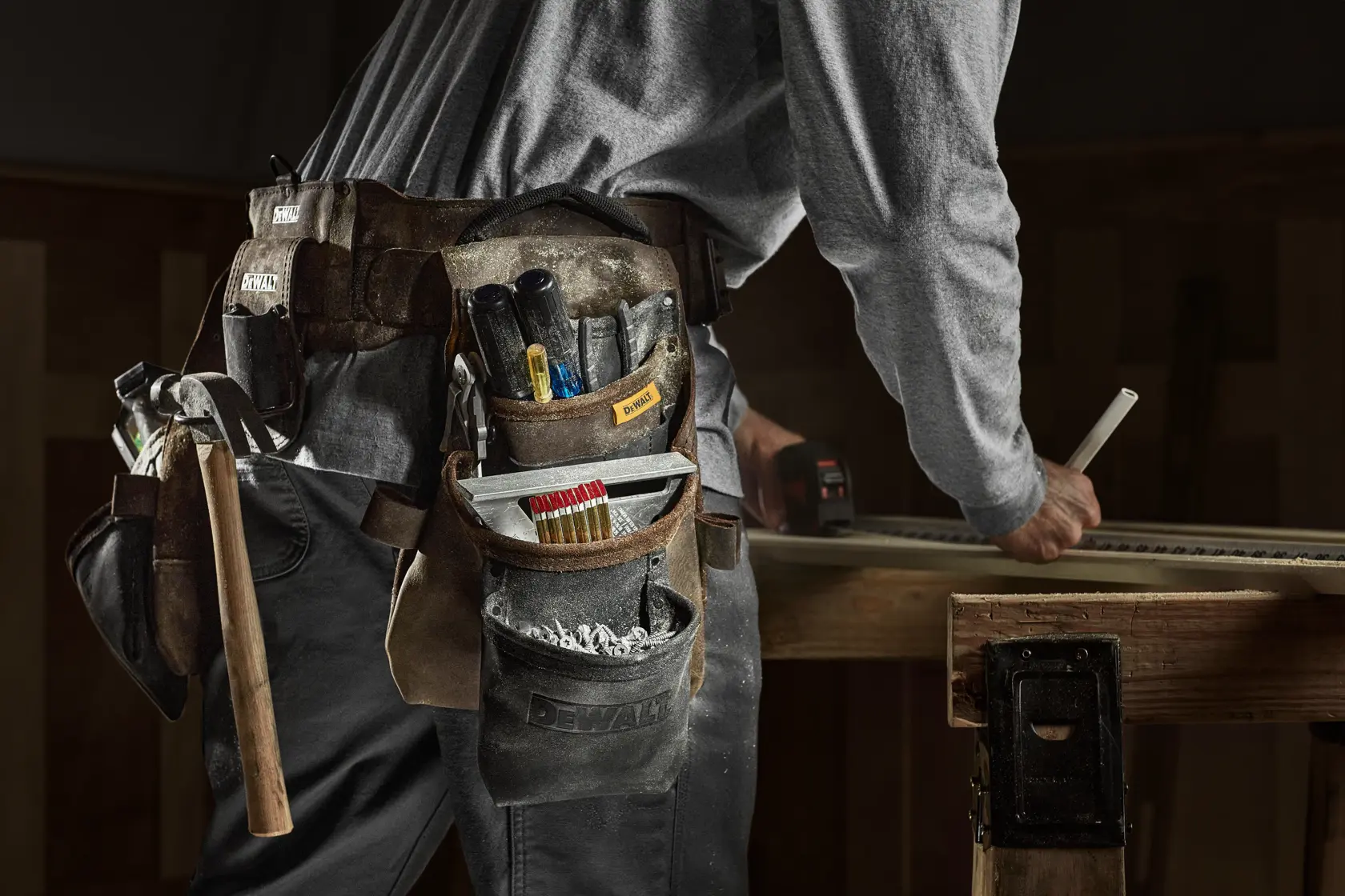 A person wearing a DEWALT tool belt with various tools such as a hammer, screwdriver, tape measure, and construction markers while working on a wooden workbench.