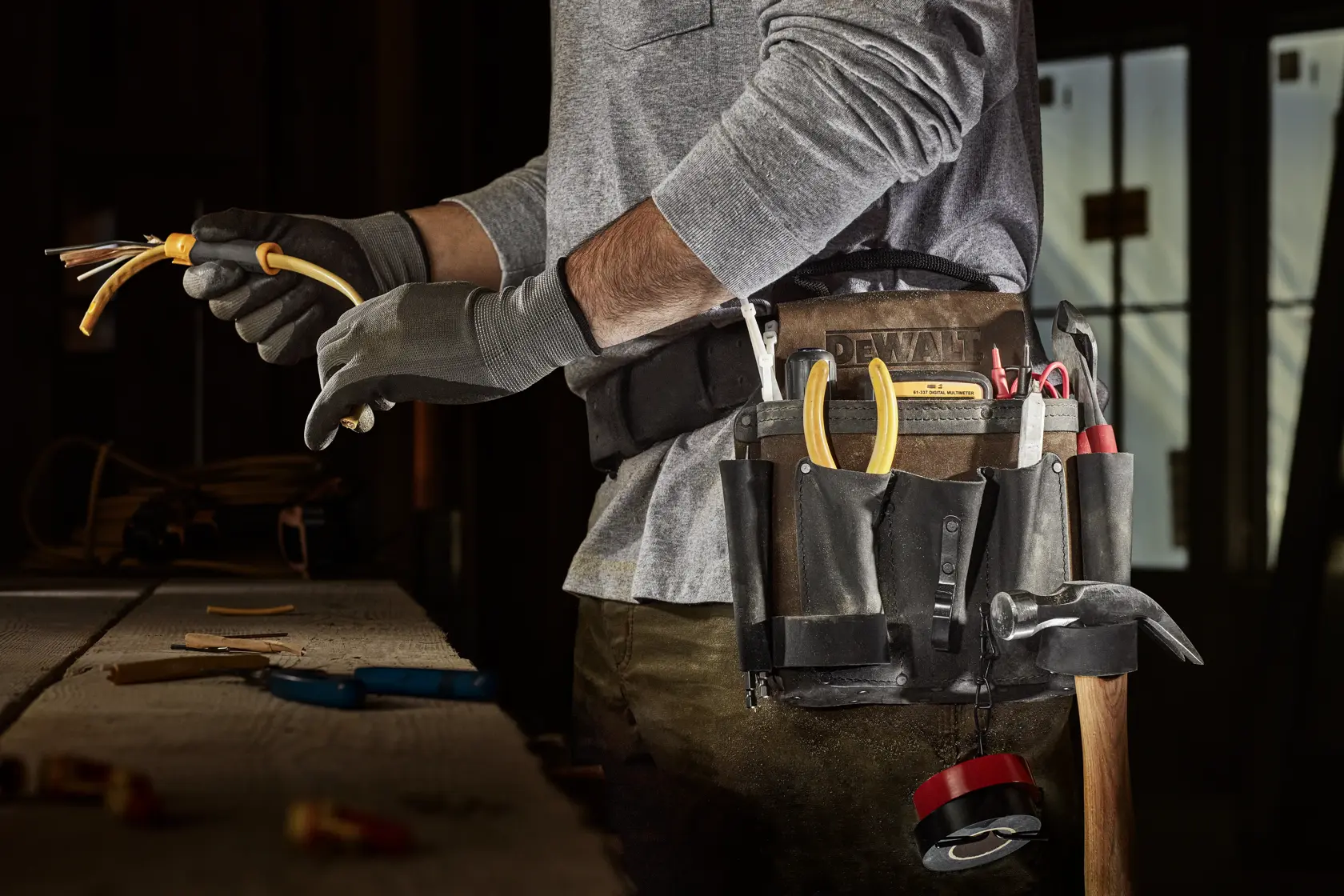 A person wearing a DEWALT tool belt with various hand tools, such as pliers, screwdriver, hammer, and a tape measure. The person is working with electrical wires on a workbench, wearing work gloves.