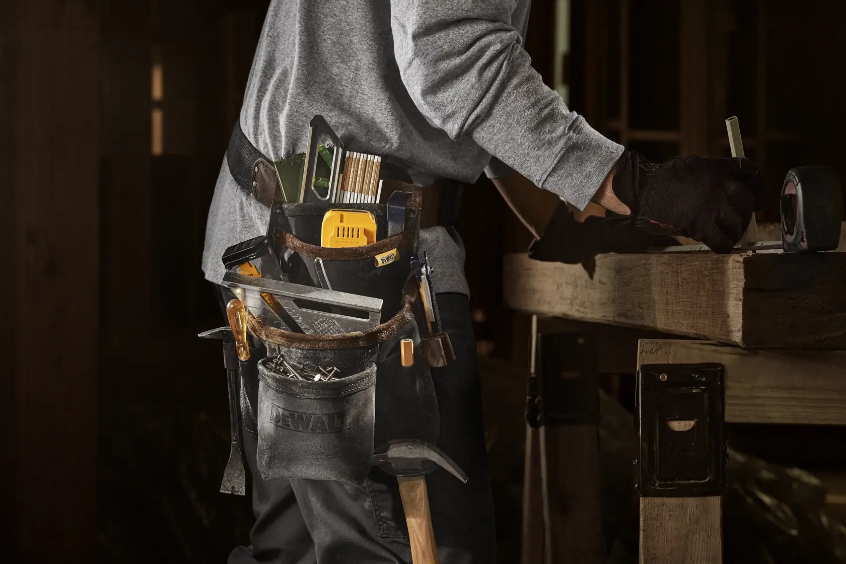 Close-up of a person wearing a DEWALT tool belt filled with various hand tools while working at a wooden workbench. The person is wearing a gray sweatshirt and black gloves.