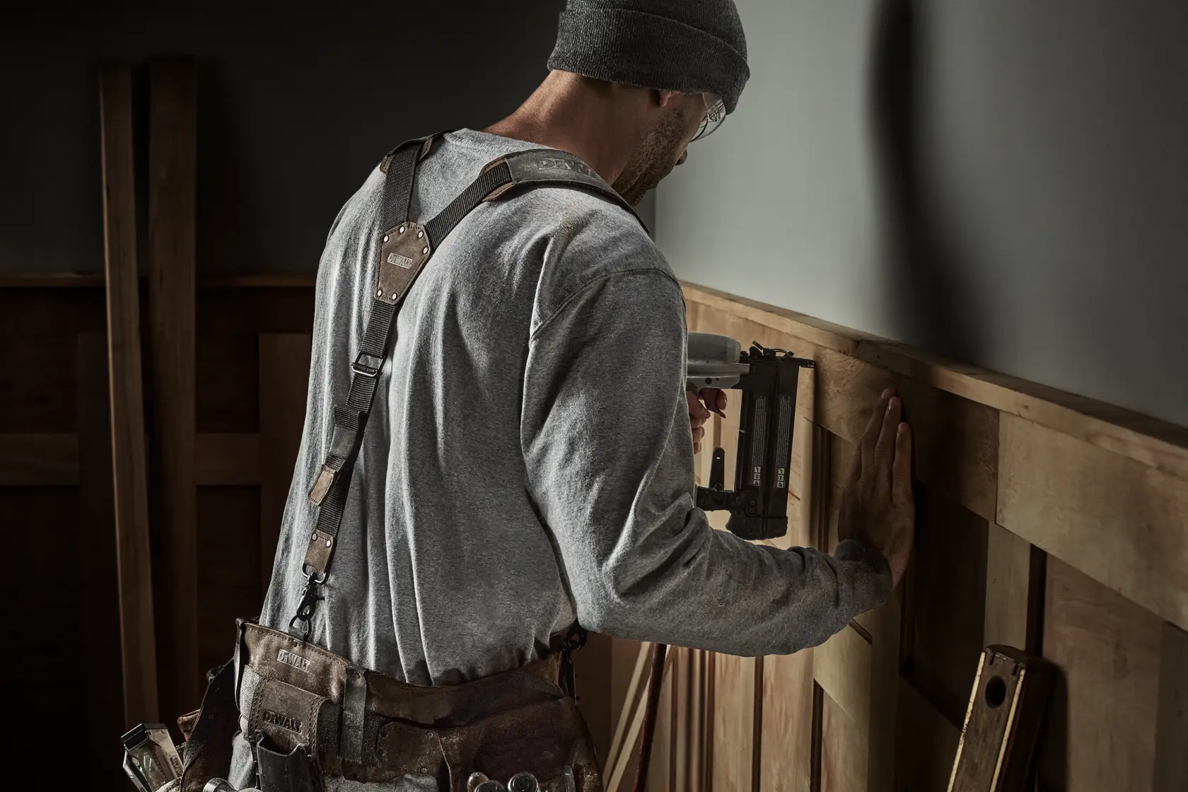 A person wearing a gray shirt and beanie is using a DEWALT nail gun to attach wooden panels to a wall. The person is also wearing a tool belt with various tools. The setting appears to be an indoor construction or renovation site.