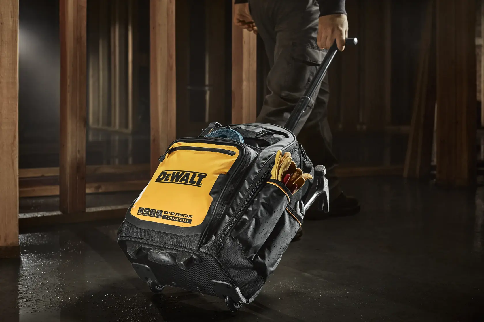 A DEWALT branded rolling tool bag with a yellow and black design, being pulled by a person in a construction setting. The bag appears to be water-resistant and contains various tools and gloves in its side pockets.