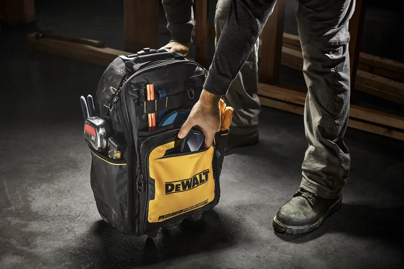 A person is holding a DEWALT tool backpack filled with various tools and accessories. The backpack is black with a yellow front pocket displaying the DEWALT logo. The scene appears to be in a workshop or construction environment.