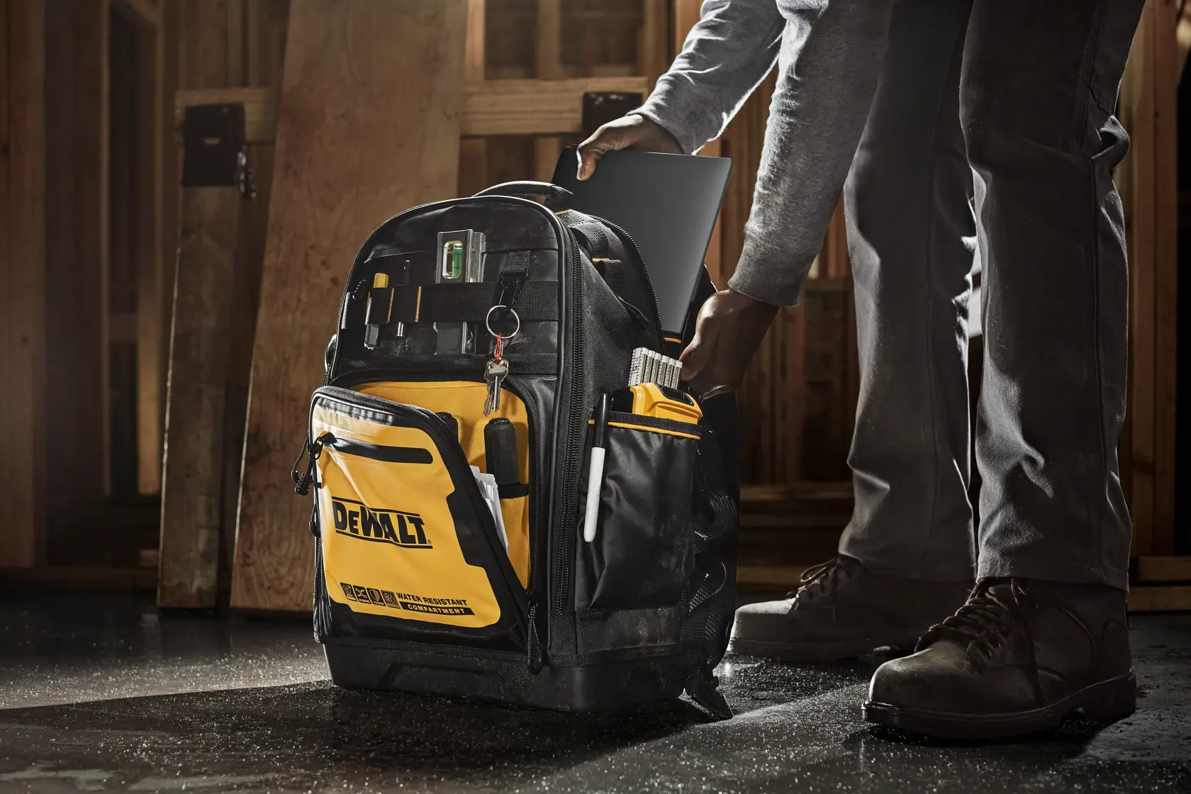A DEWALT tool backpack with multiple compartments and organized tools, being used at a construction site. A person is placing a tablet into the backpack.