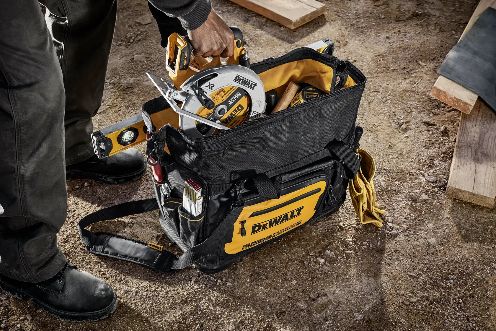 A person is placing a DEWALT circular saw into a black and yellow DEWALT tool bag at a construction site. The bag contains various tools including a spirit level and is set on a dirt ground with wooden planks nearby.