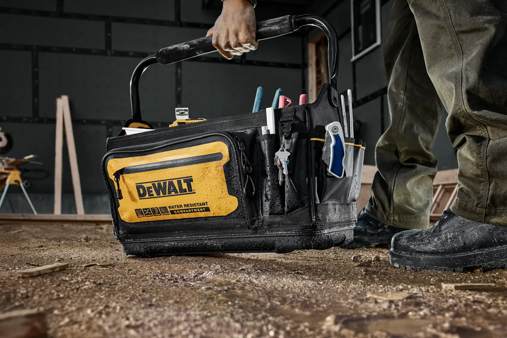 A person holding a DEWALT water resistant tool bag filled with various tools, standing on a dusty workshop floor.