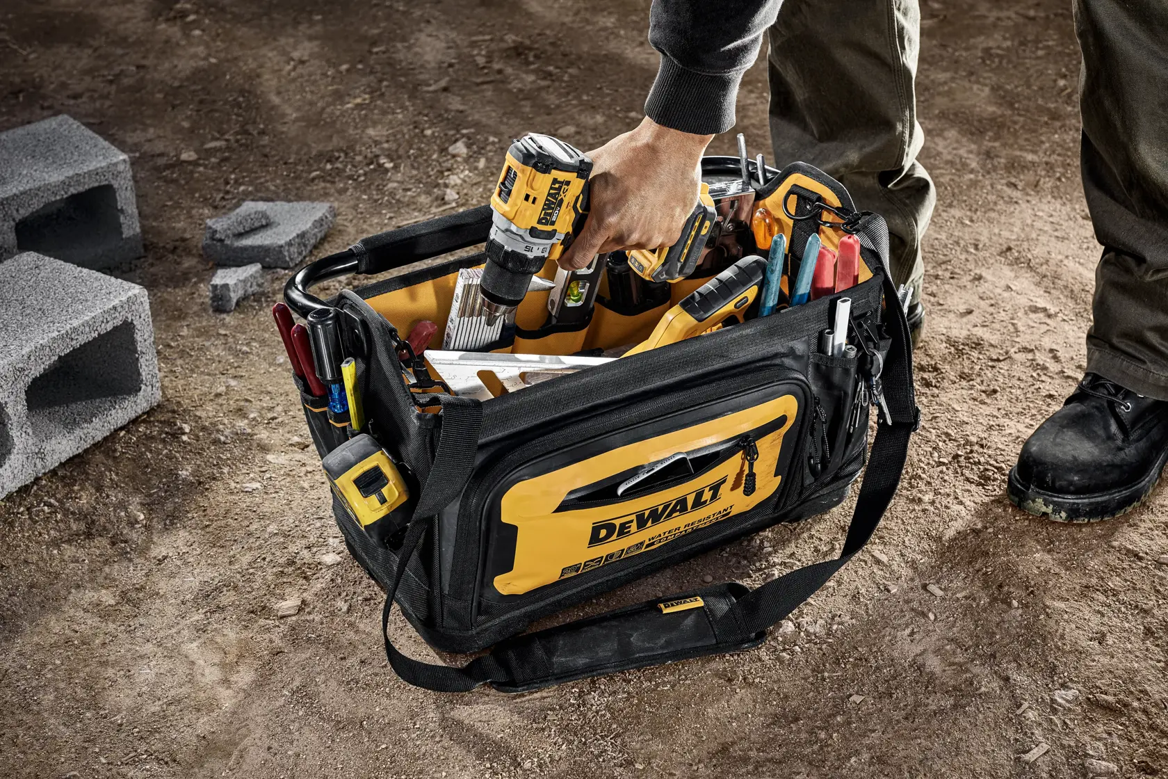 A black and yellow DEWALT tool bag filled with various tools is placed on the ground. A person is reaching into the bag and holding a cordless DEWALT drill. The bag is surrounded by construction materials, and the person is wearing work boots.