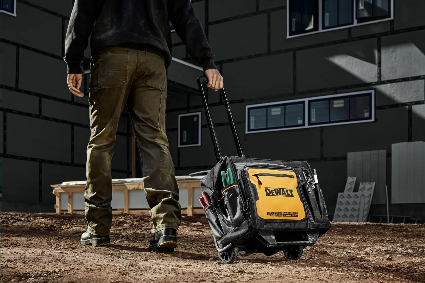 A person is walking outdoors on a construction site, pulling a DEWALT branded rolling tool bag filled with tools. The background shows a partially constructed building.