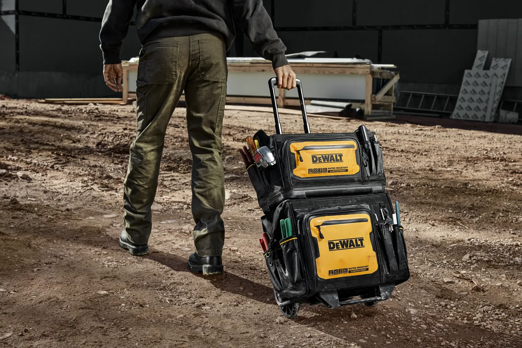 A person is walking on a construction site, pulling a DEWALT rolling tool bag filled with various tools. The tool bag is black with yellow DEWALT branding and has multiple pockets and compartments.