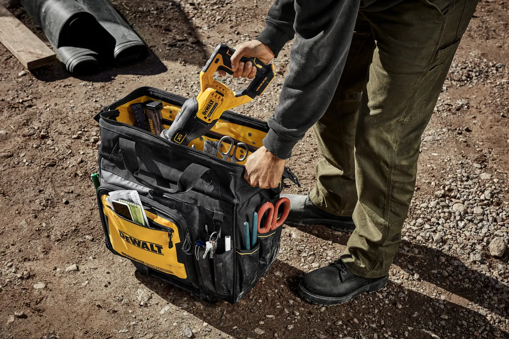 A person standing outdoors on a gravel surface is holding a yellow and black DEWALT power tool above a large DEWALT-branded tool bag. The bag is filled with various tools and accessories, including scissors and measuring instruments in exterior pockets.
