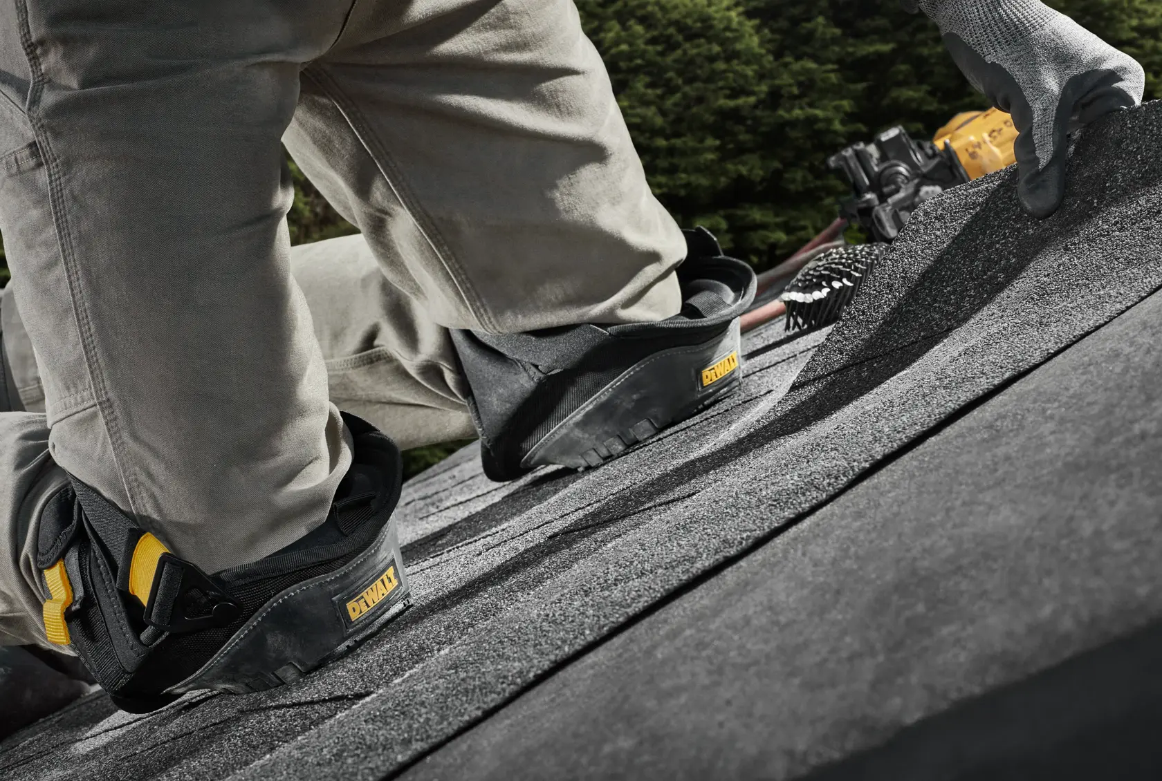 A person wearing DEWALT knee pads is kneeling on a shingle roof, working outdoors. The focus is on the protective knee pads and the textured surface of the roof.