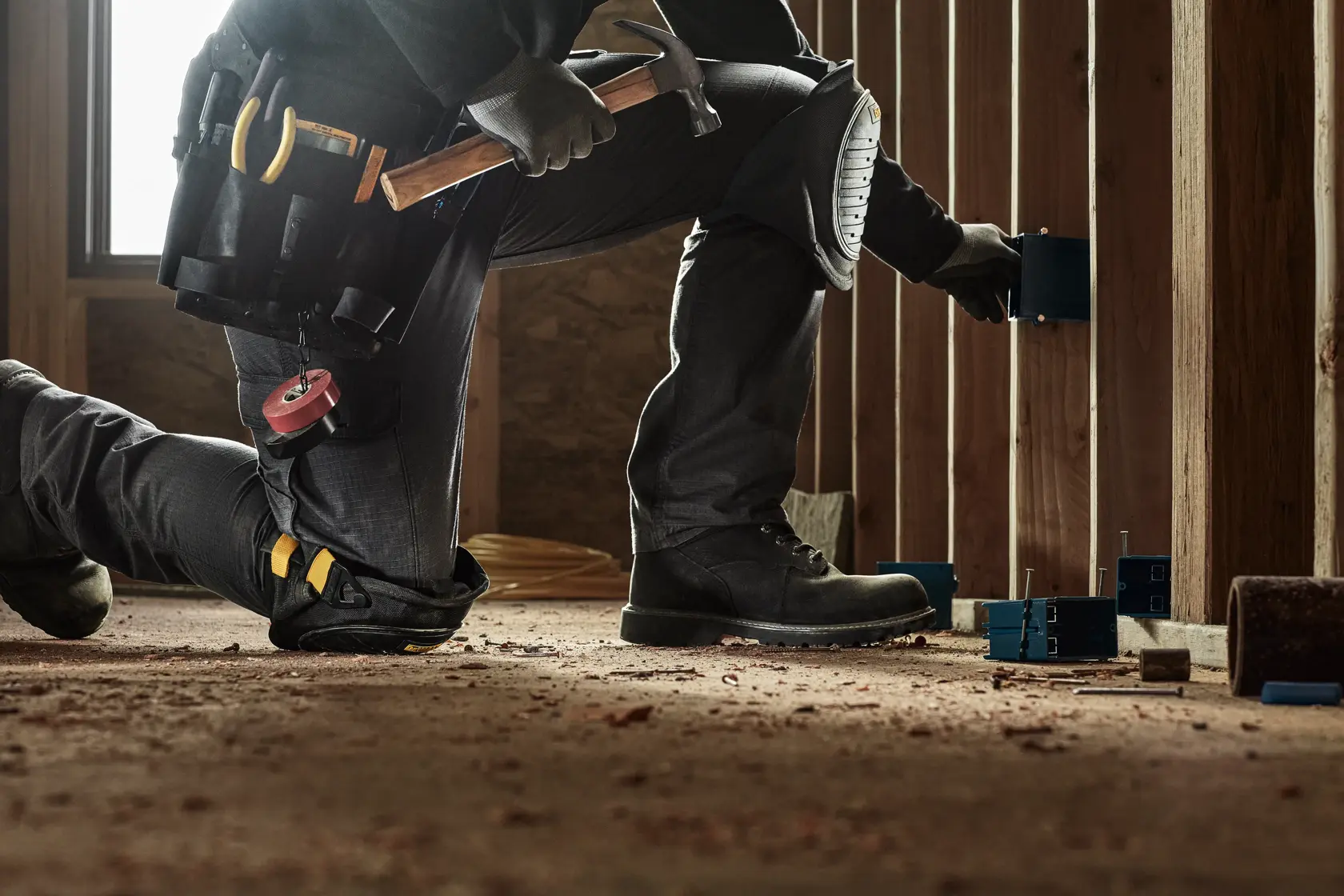 A person wearing work boots, kneepads, and a tool belt with various tools, including a hammer, is kneeling on a construction site floor and installing electrical boxes into wooden wall studs.