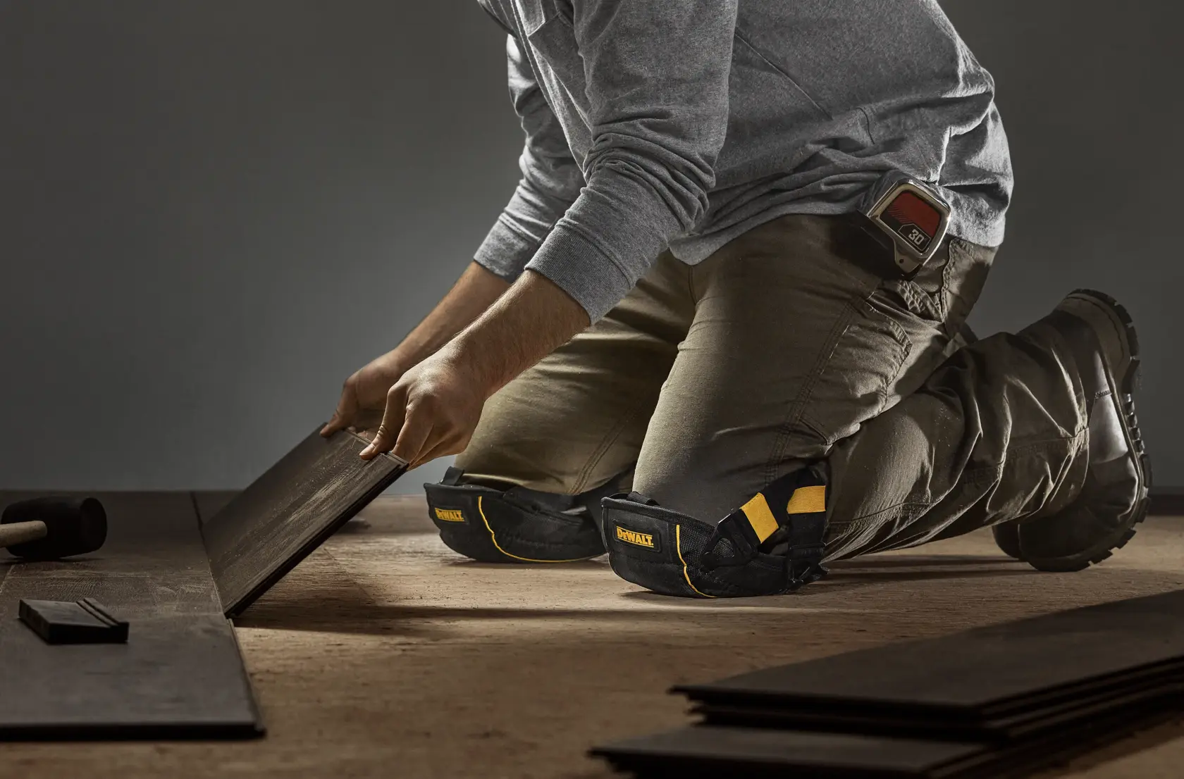A person wearing DEWALT knee pads is kneeling on the floor while installing dark wooden flooring panels. The individual is dressed in work attire and boots, with tools visible around.