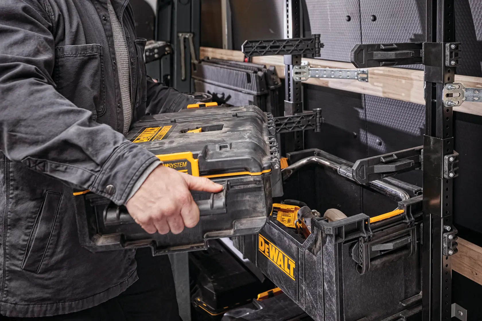 A person places a DEWALT ToughSystem toolbox onto a storage rack inside a vehicle. Several DEWALT storage boxes and tools are visible, and the rack is mounted to the wall for secure organization.