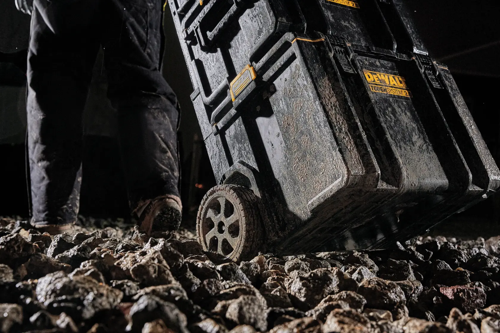 A close-up view of a DEWALT ToughSystem rolling tool storage box being pulled over rough, rocky ground. The box and wheels are muddy, emphasizing durability and rugged use.