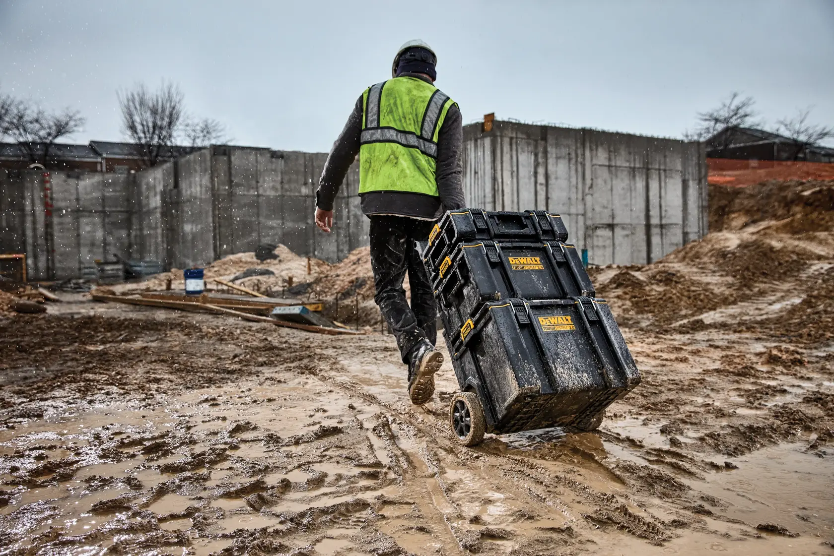 A worker with a green safety vest and helmet pulls a black DEWALT rolling tool storage box across a muddy construction site, with concrete walls and building materials visible in the background.