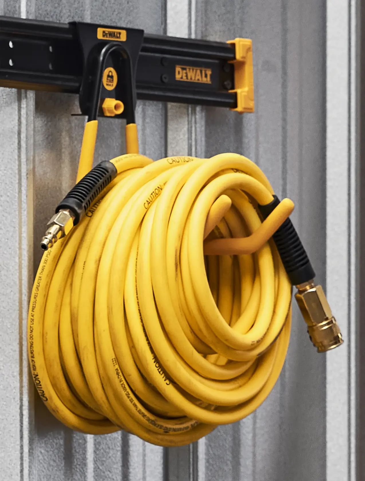 A yellow DEWALT hose with metal connectors is neatly coiled and hung on a DEWALT branded wall-mounted rack against a corrugated metal wall.