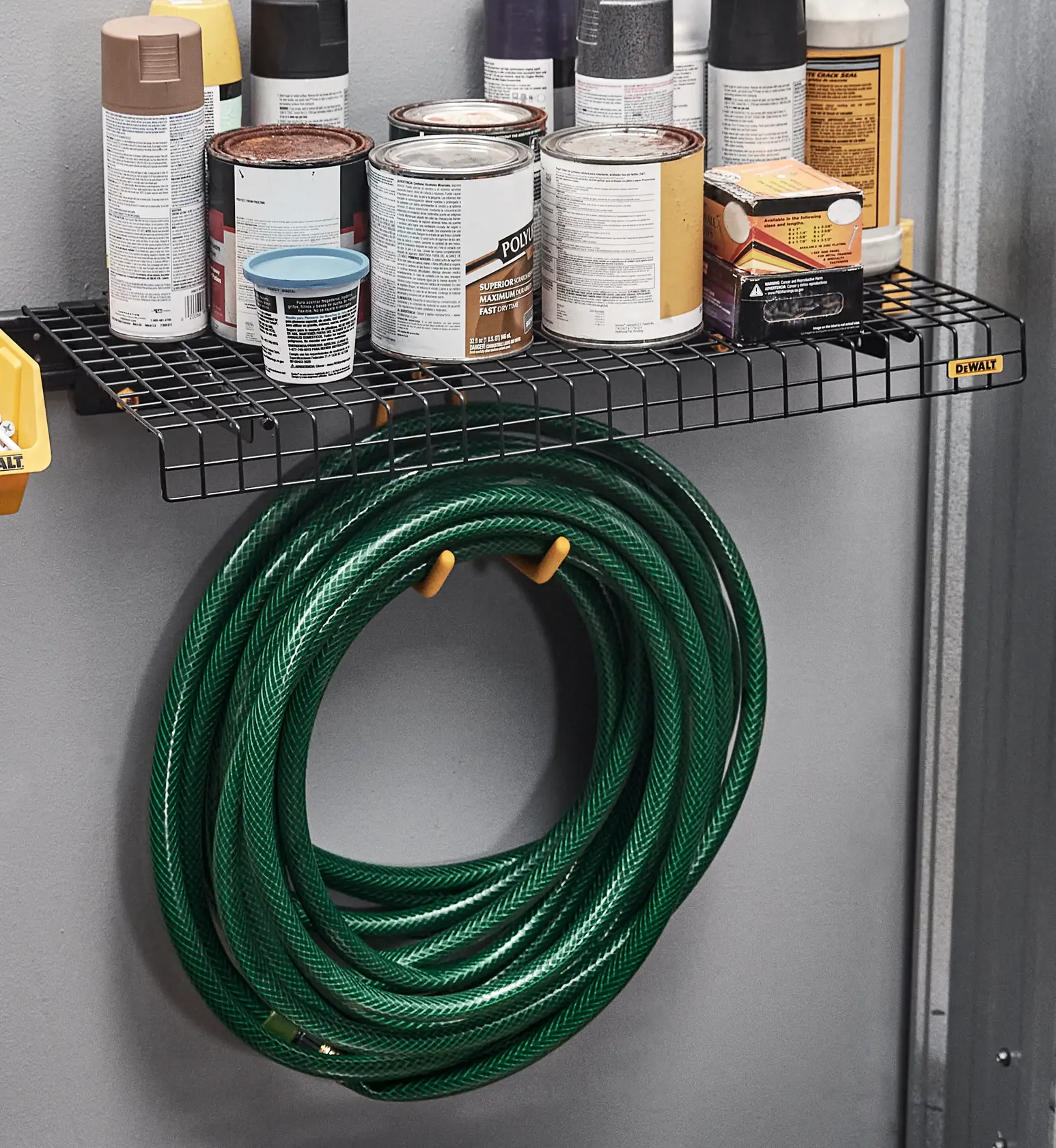 A DEWALT branded wire shelf mounted on a wall, holding cans, spray bottles, and other containers, with a green garden hose neatly coiled and hung underneath the shelf using two yellow hooks.