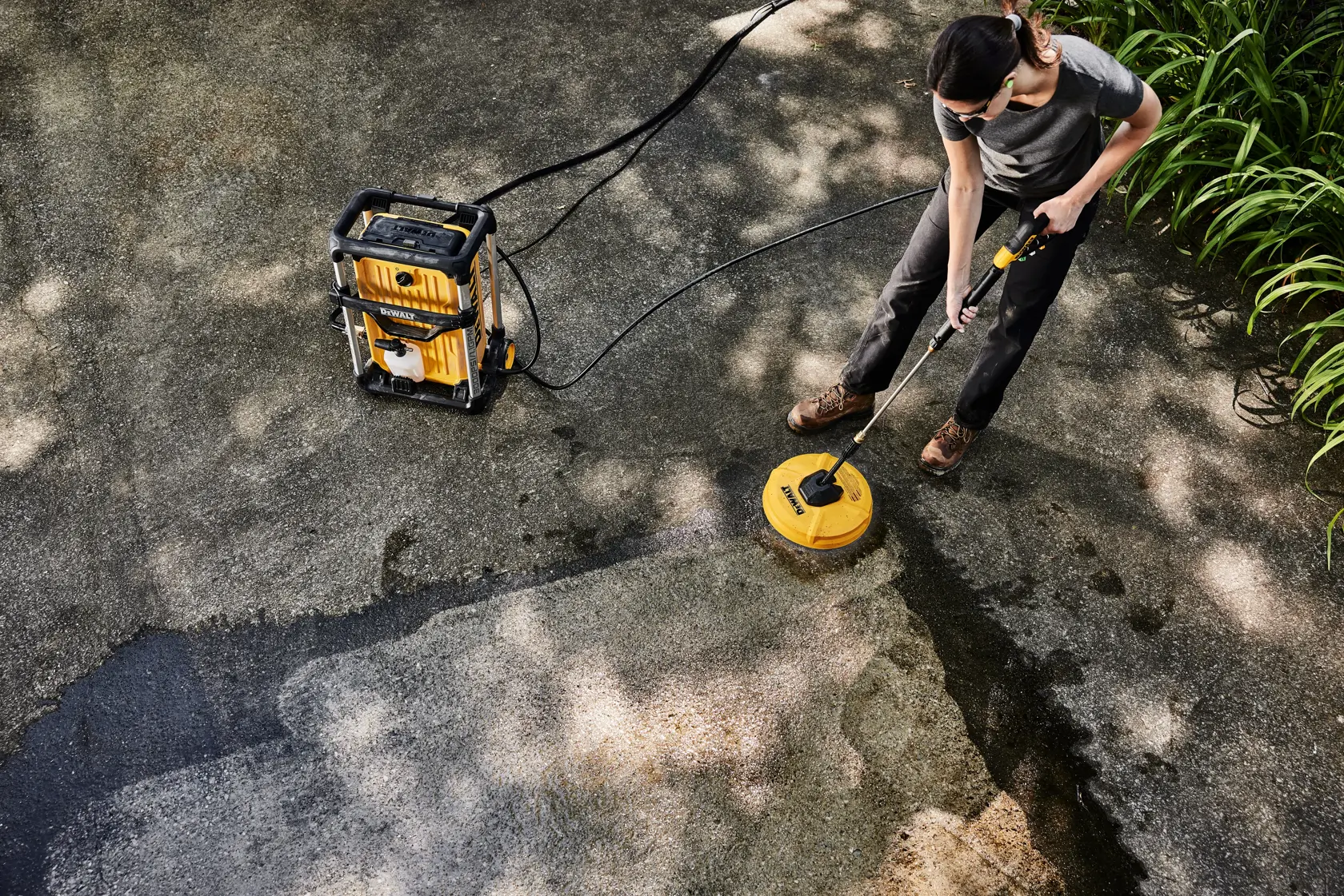 A person uses a DEWALT pressure washer and surface cleaner attachment to clean a concrete driveway. The equipment has a yellow and black color scheme and is connected with hoses. The driveway shows a noticeable clean section where the surface cleaner was used, and there are plants on the side.
