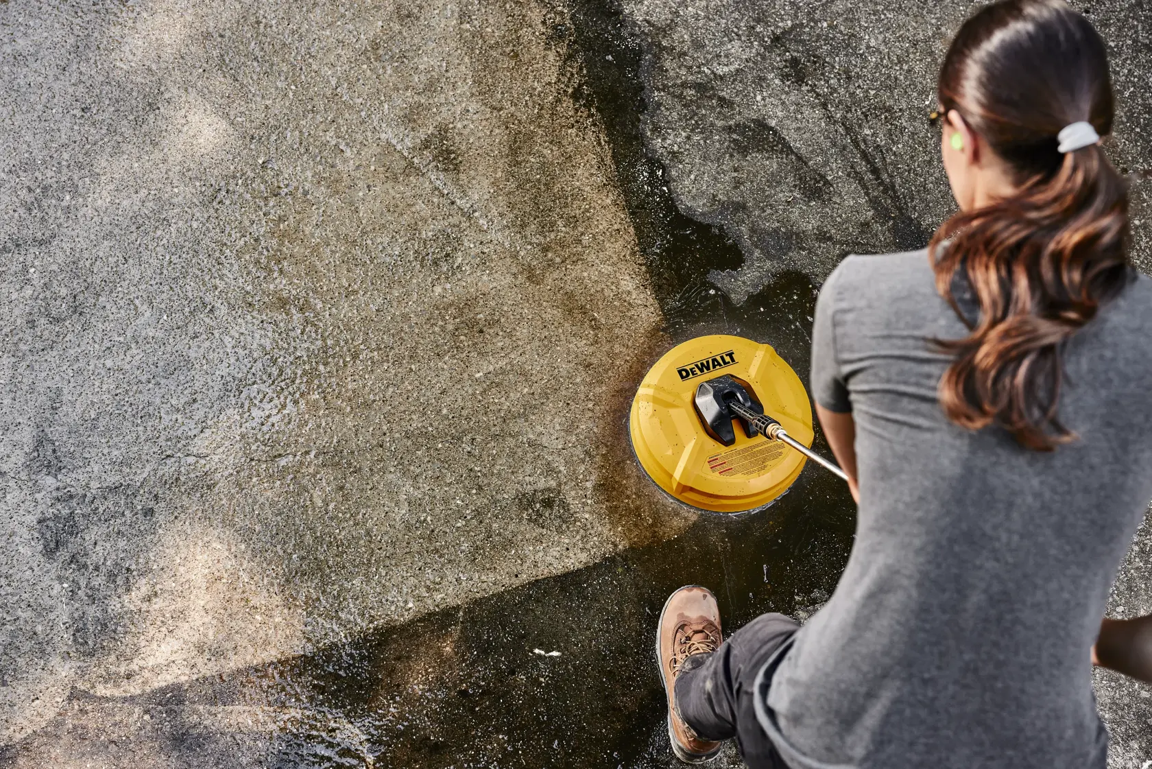 A person is using a DEWALT surface cleaner attachment to clean a concrete outdoor surface. The yellow cleaning tool is making a noticeable difference between the cleaned and uncleaned areas.