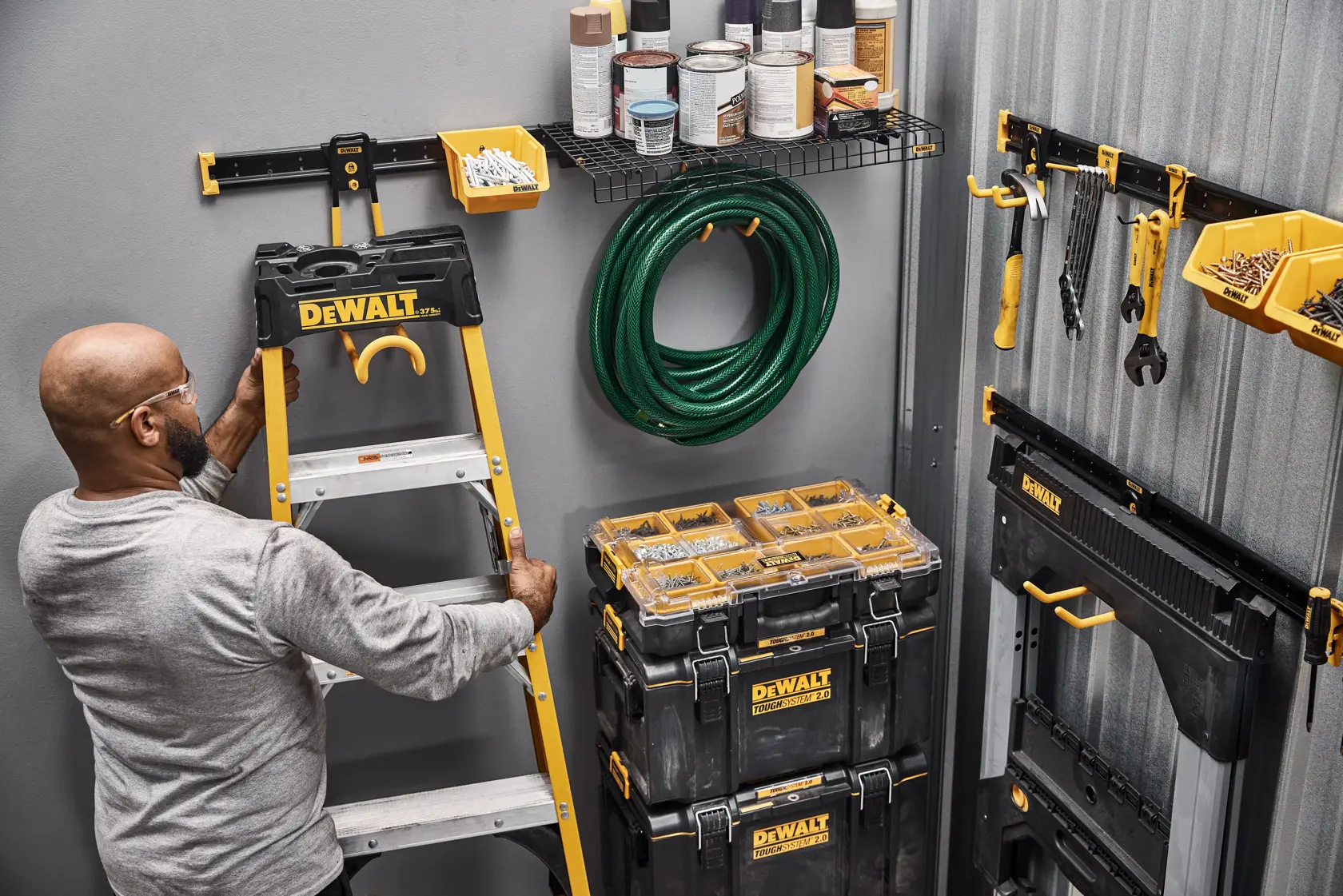 A person organizing a workspace with DEWALT storage products, including a ladder, toolboxes, wall-mounted bins, hand tools, a garden hose, and shelves with paint cans.