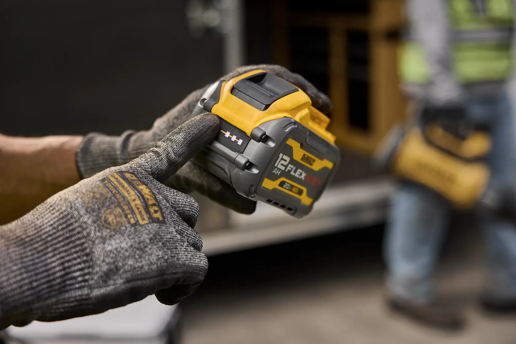 Gloved hand holds a DEWALT FLEXVOLT 12Ah battery for power tools on a worksite.
