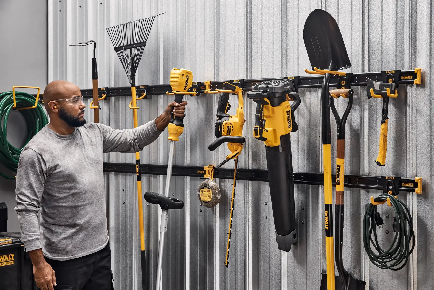 A person standing in a garage or workshop next to a DEWALT wall-mounted storage system holding a yellow and black garden tool. Various DEWALT tools and gardening implements, including a leaf blower, shovel, rake, and hose, are neatly organized on the storage rack attached to a metal wall.