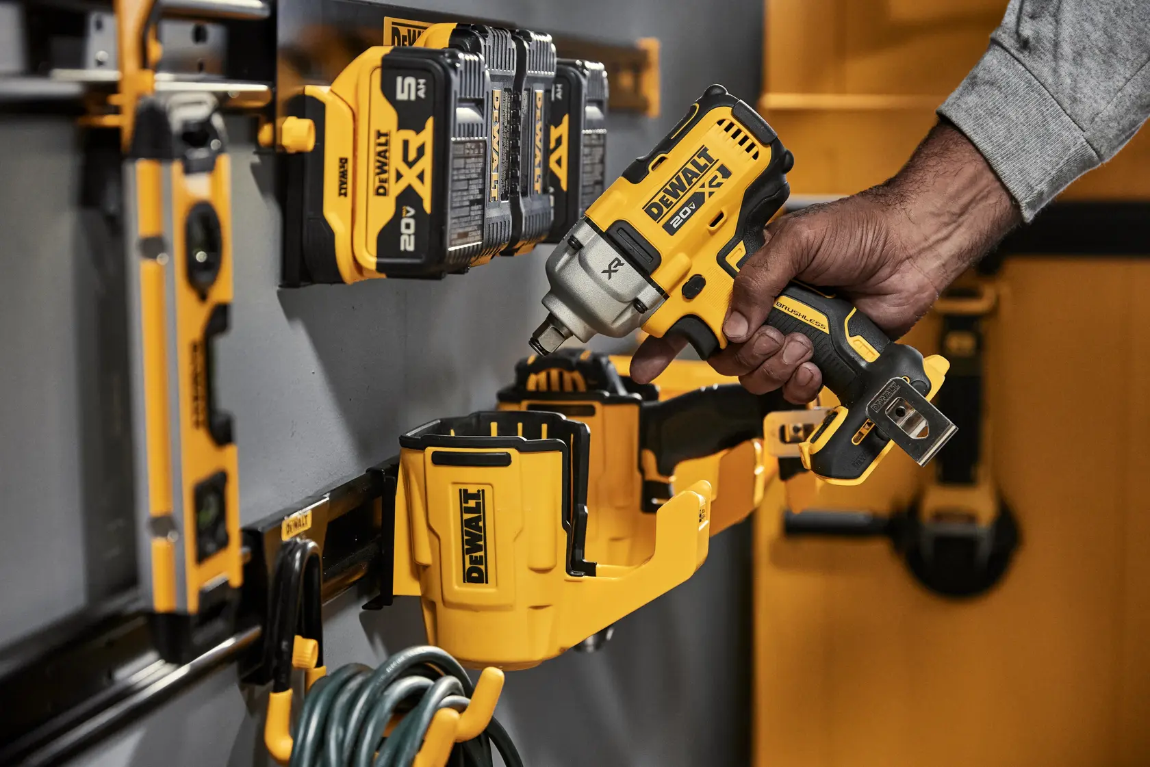 A close-up of a DEWALT wall-mounted organizer holding several yellow and black DEWALT XR 20V batteries and a DEWALT power tool being placed by a person's hand. Other DEWALT accessories and tools, including a level and cable, are visible on the wall.