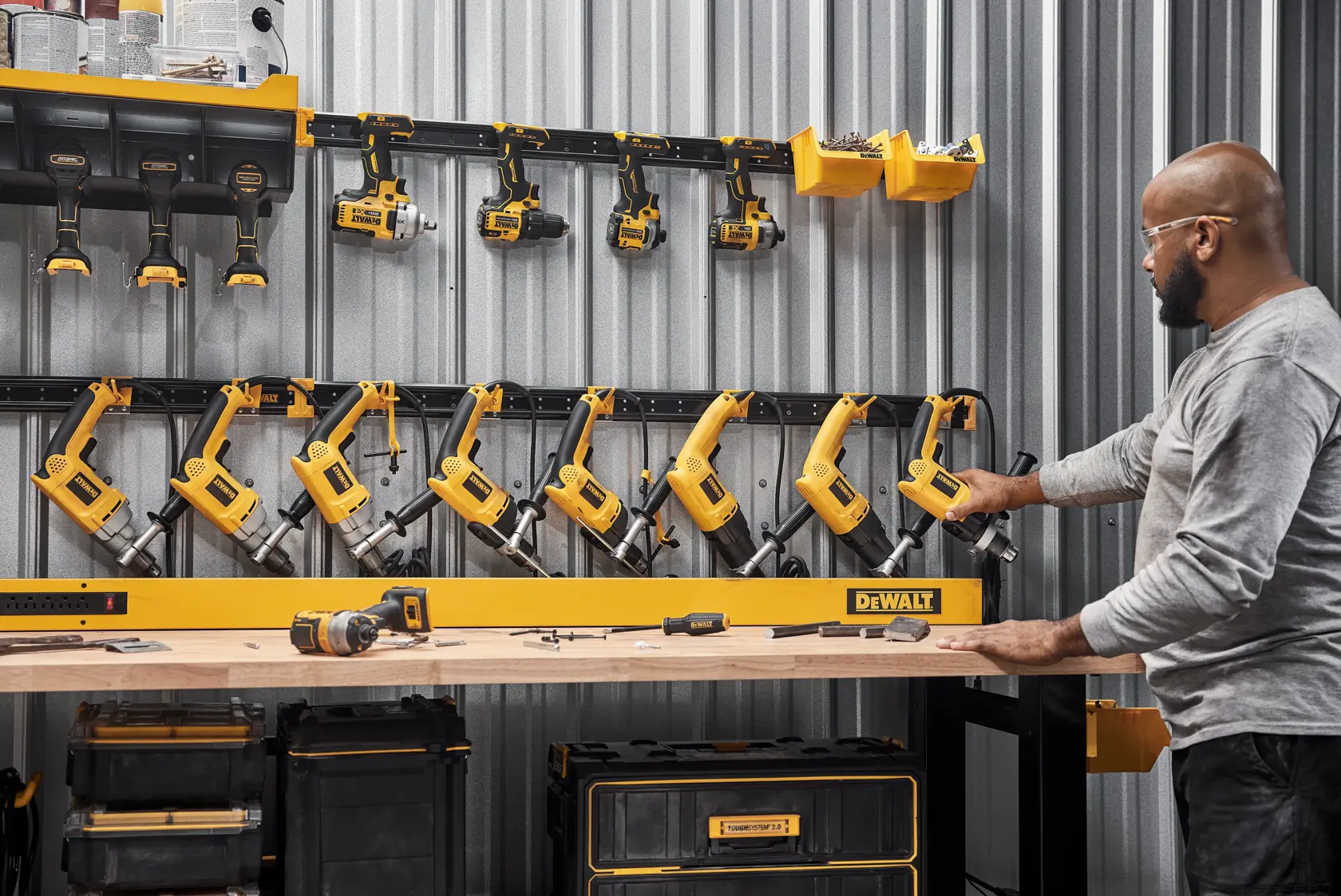 A person is organizing yellow DEWALT power tools on a wall-mounted storage rack in a workshop. The wall is made of corrugated metal, and multiple tools and accessories are neatly arranged, including drills and bins. A wooden workbench with various tools is visible in the foreground.