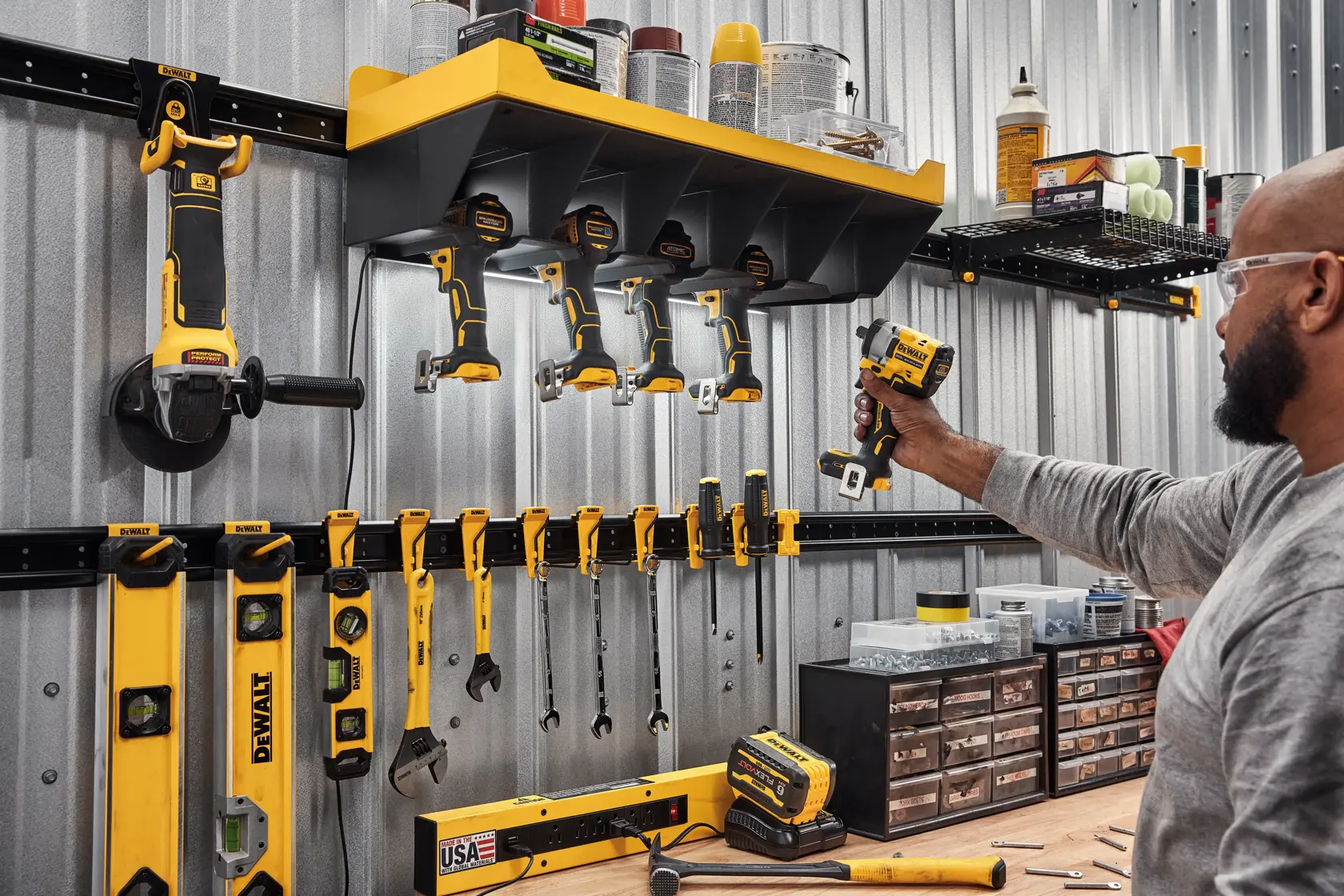 A well-organized workshop tool wall featuring several yellow and black DEWALT power tools, hand tools, and levels. Tools are neatly hung on a rail system and shelves, with various supplies and hardware stored above and below. A person in a gray shirt, with their face blurred, is holding a cordless power tool.