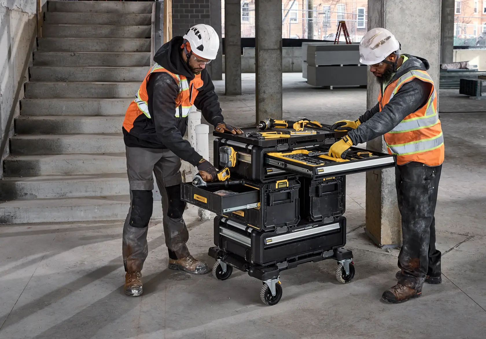 Two construction workers wearing safety helmets and vests are using a DWST08550 DEWALT rolling tool storage cart in a building under construction. The cart has multiple drawers and compartments open, showing various DEWALT tools placed on top and inside. The cart is positioned on wheels for mobility.