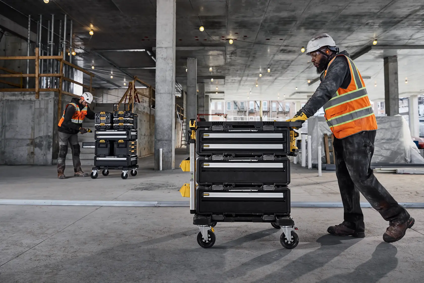 Two workers in safety gear and orange vests use DEWALT portable tool storage carts with multiple drawers on wheels inside a large construction site with concrete columns and overhead lights.