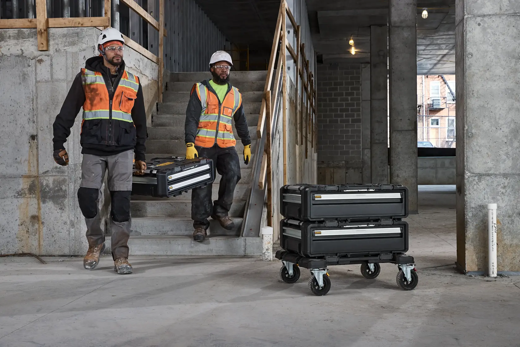 Two construction workers wearing safety vests and helmets carry DEWALT tool cases down a concrete staircase at a job site. Nearby, more stacked DEWALT tool cases are placed on a rolling cart. The setting includes unfinished concrete walls and structural elements.